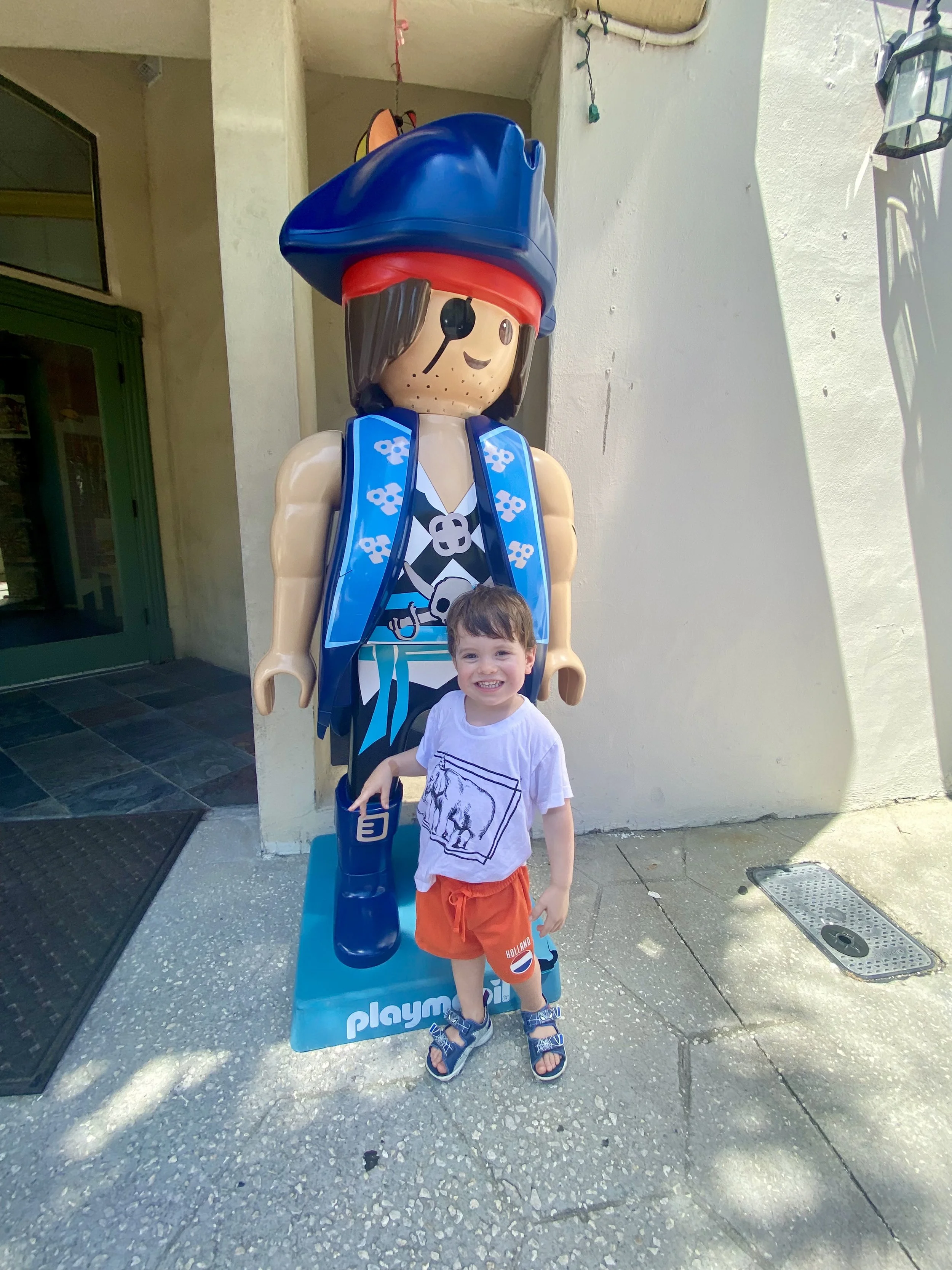 A young boy smiling and standing in front of a pirate-themed sculpture at Playmobil land. The sculpture is a large cartoon pirate with a blue hat, eyepatch, and a blue vest with skull and crossbones. The boy is wearing a white t-shirt with a wolf print, orange shorts, and sandals.