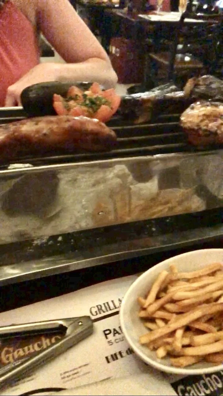 Grilled food being cooked on an outdoor grill, with a bowl of French fries and a menu in the foreground.