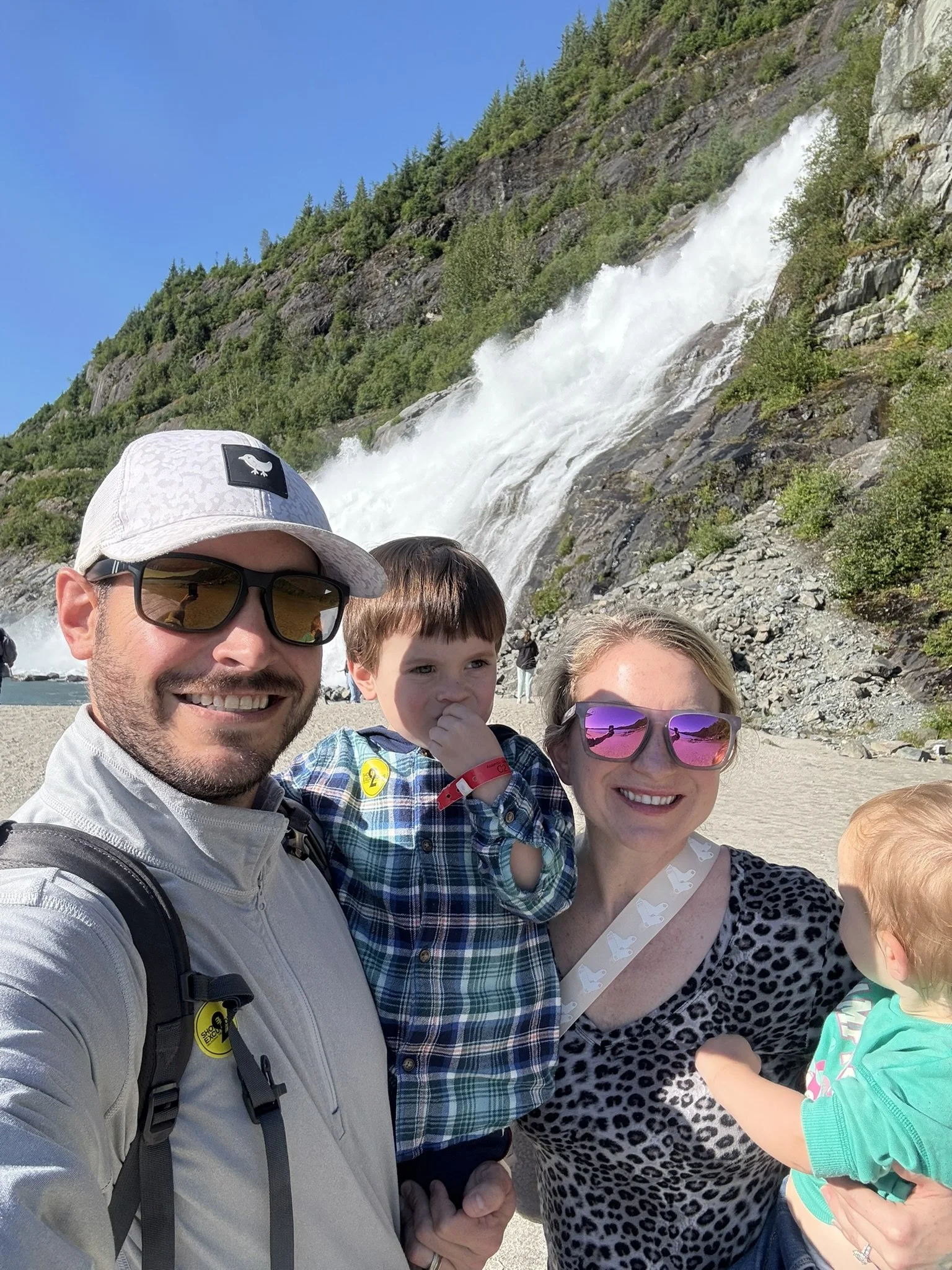 A smiling family of four taking a selfie in front of a waterfall and mountain landscape on a sunny day.