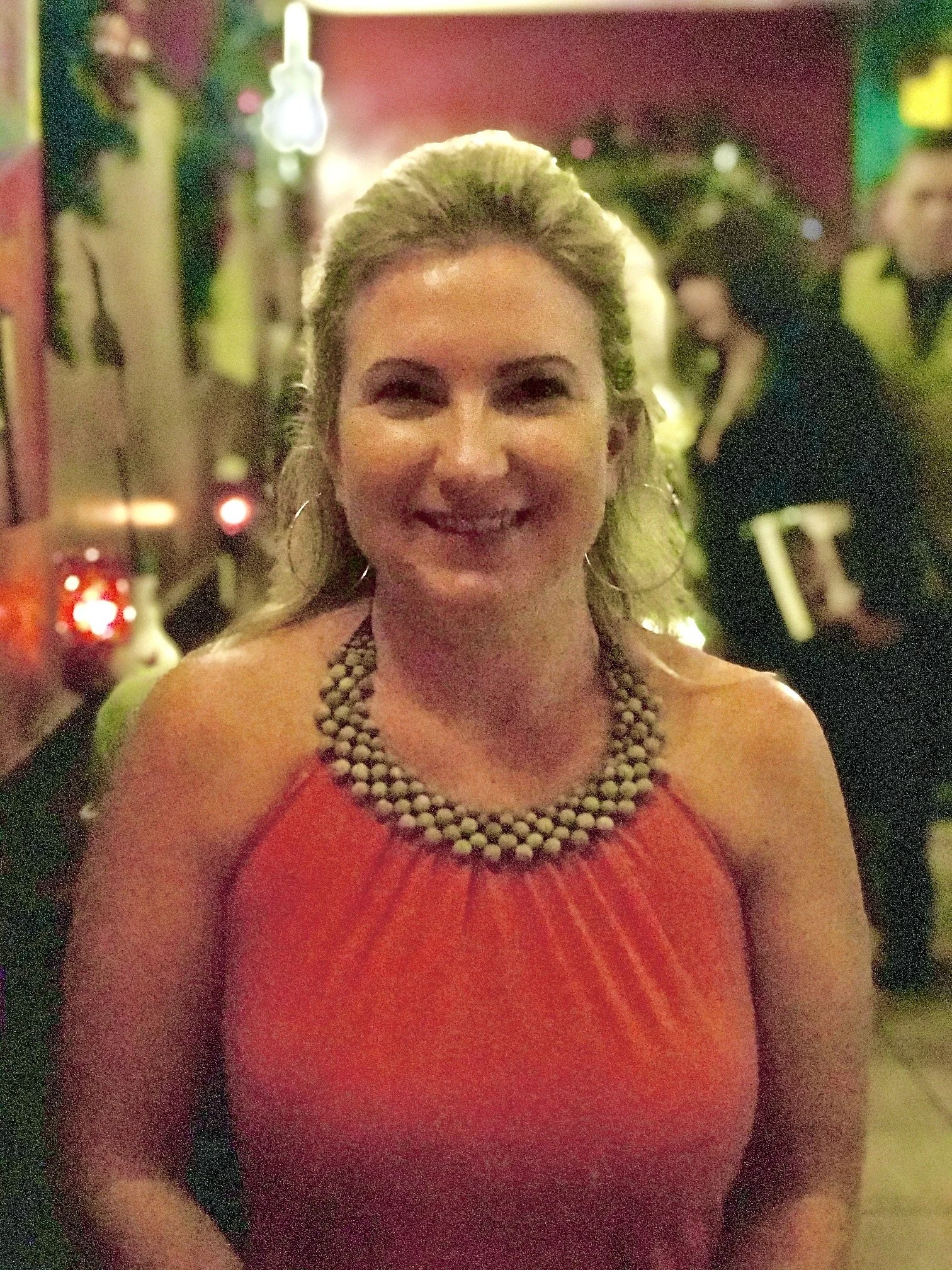 A woman taking a selfie in a dimly lit, colorful party or nightclub scene, wearing a red dress with a statement necklace, smiling.