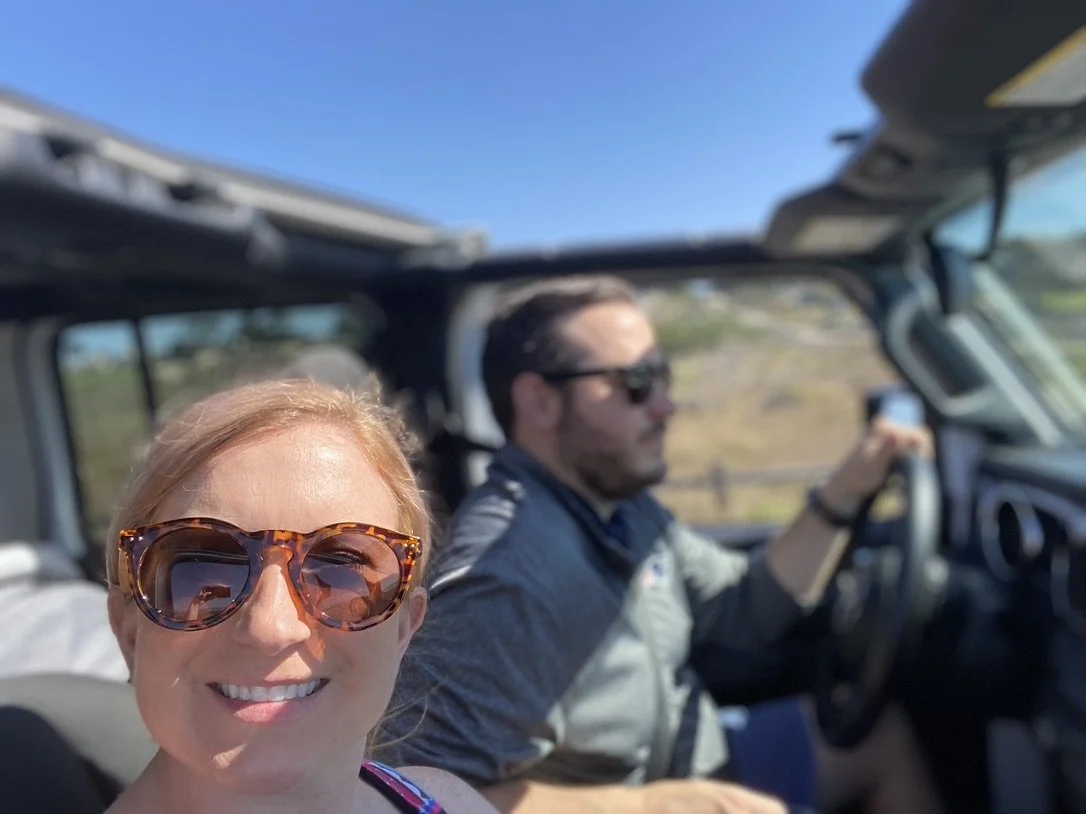 A woman with blonde hair and tortoiseshell sunglasses smiling in the foreground, a man with dark hair and sunglasses driving a vehicle in the background, both under a blue sky.