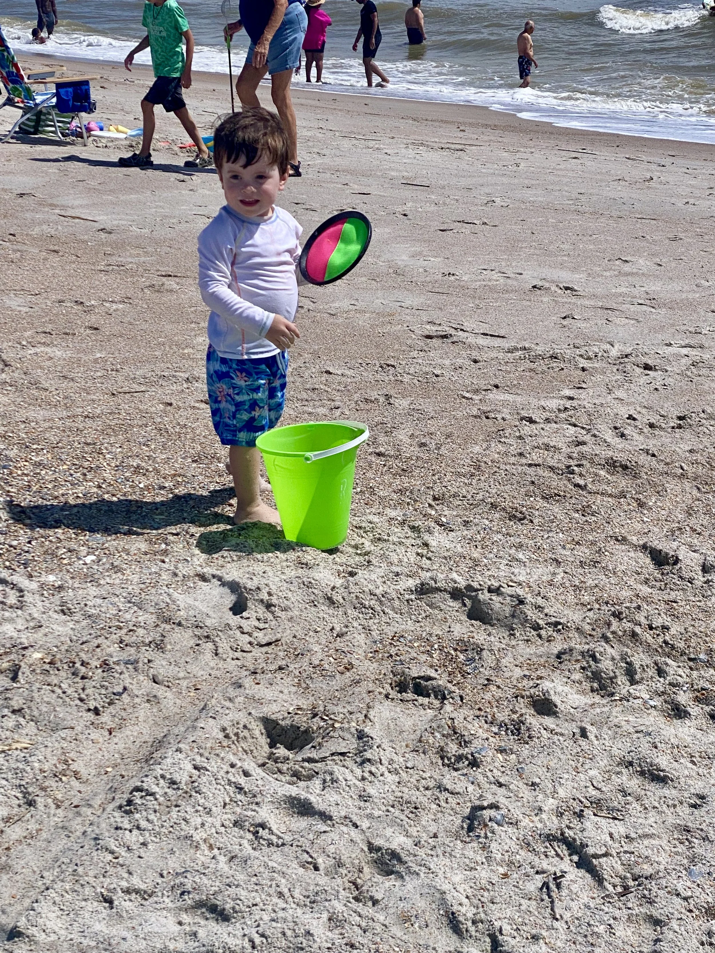 Young boy on the beach holding a colorful Frisbee next to a green bucket, with other beachgoers and the ocean in the background.