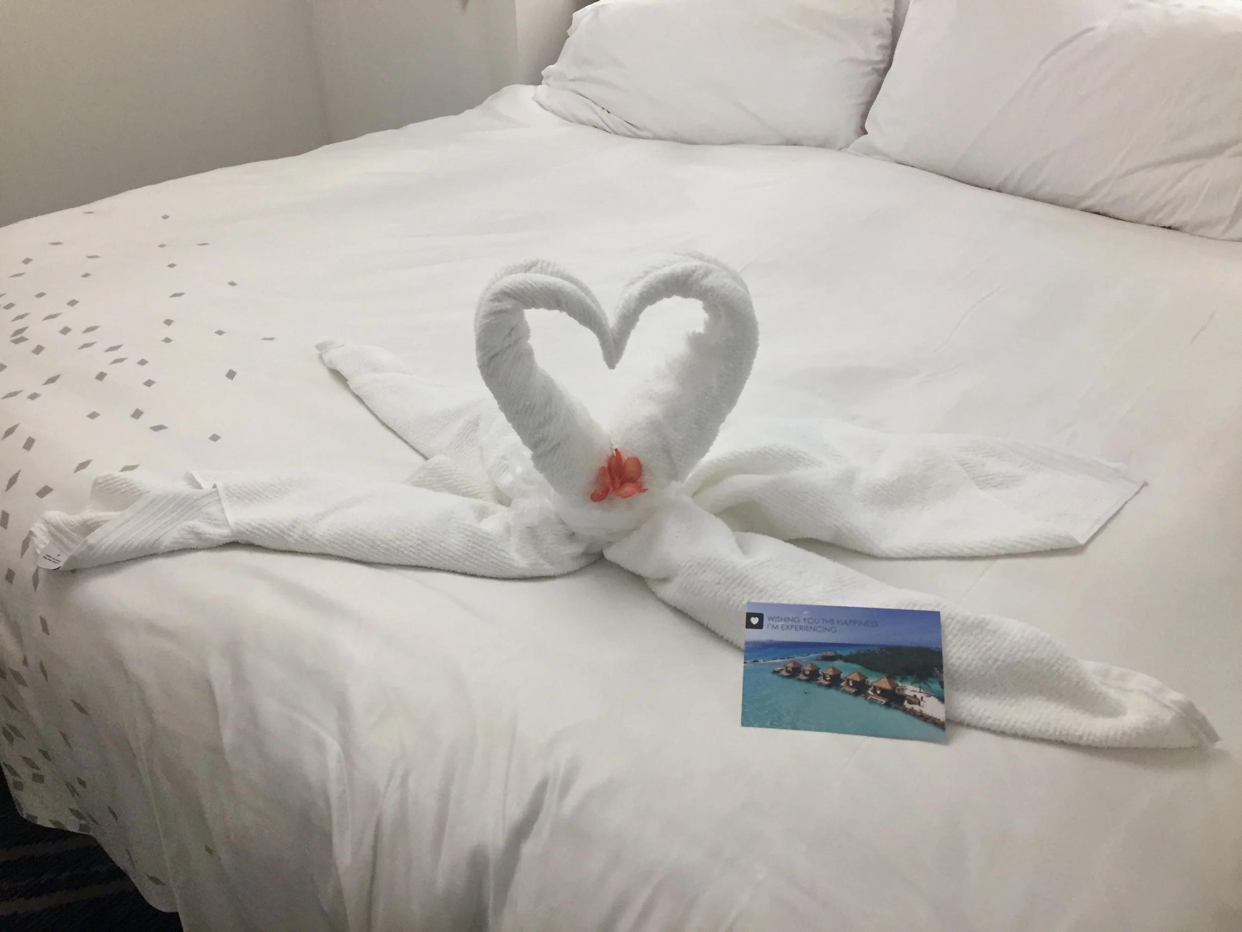A bed with white linens and pillows, decorated with two towel swans forming a heart shape. There is a colorful postcard featuring overwater bungalows on a tropical beach at the foot of the bed.