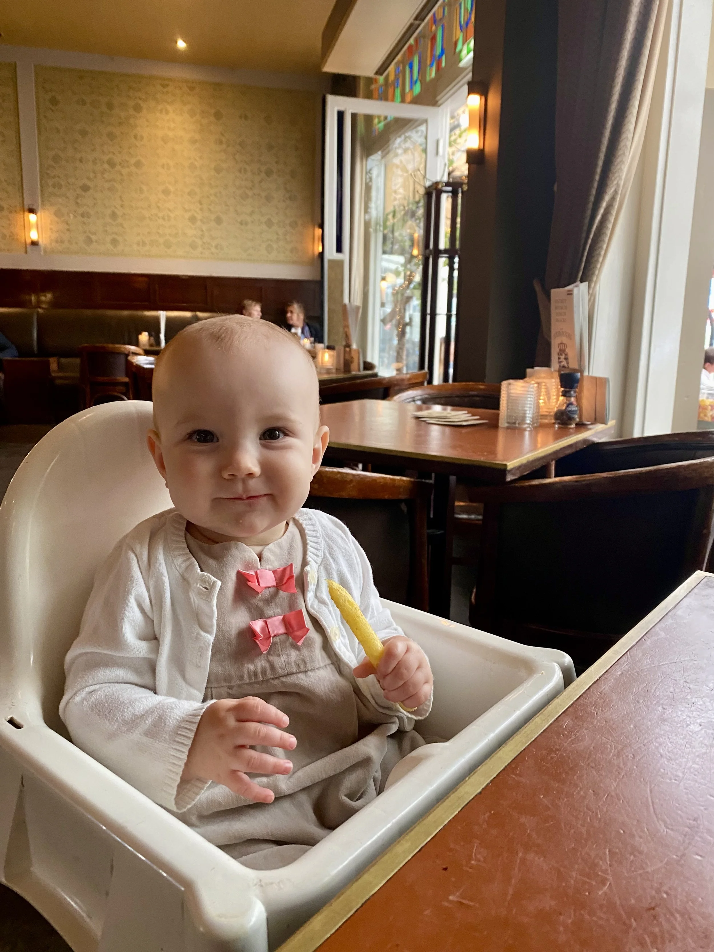 A smiling baby girl sitting in a high chair at a restaurant, holding a French fry, with indoor tables and a window with stained glass in the background.
