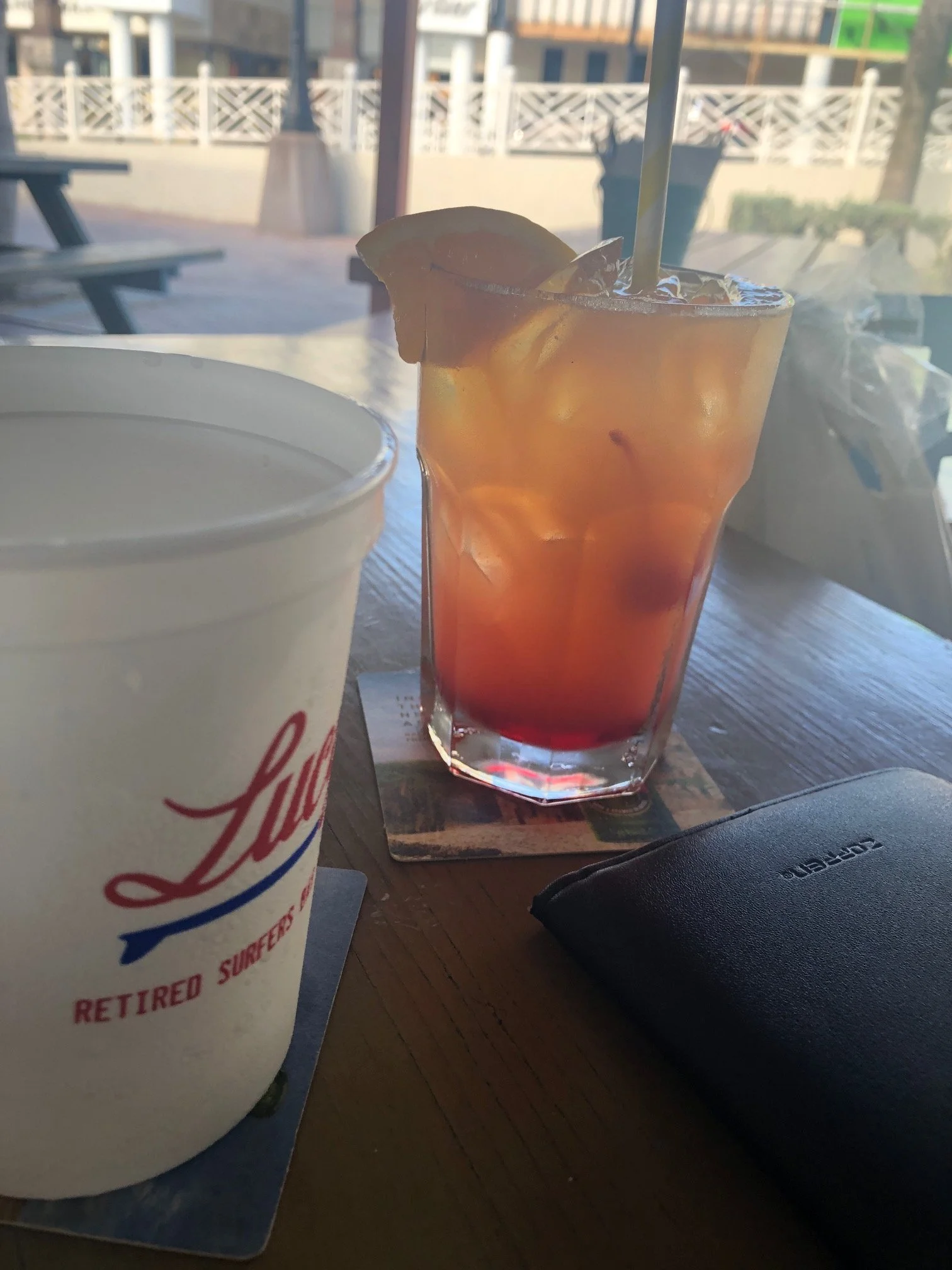 A table with a cup of iced tea or soda, a paper cup with Jason's Deli logo, and a black wallet. Outside view with a wooden fence, trees, and some construction or buildings in the background.