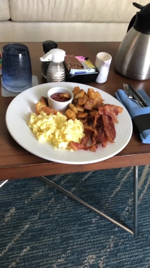 Plate of scrambled eggs, bacon, crispy hash browns, and a small cup of ketchup on a wooden table.