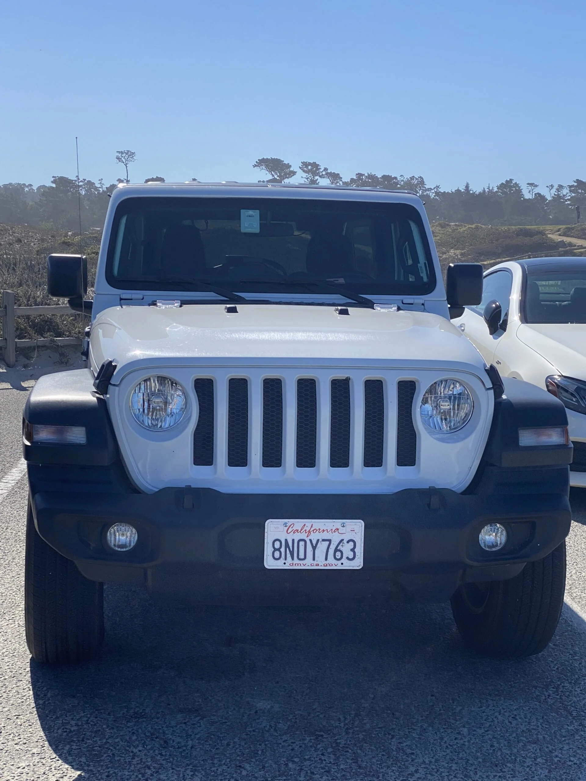 Front view of a white Jeep with a California license plate number 8NOY763 parked on a paved surface, with a clear blue sky and trees in the background.