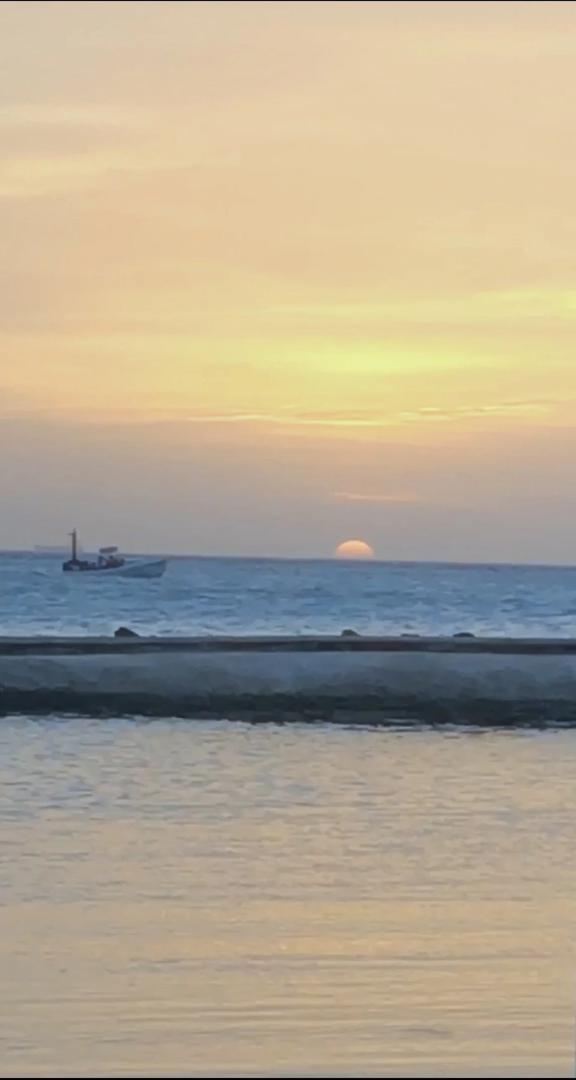 Sunset over the ocean with a boat sailing in the distance and a shoreline in the foreground.