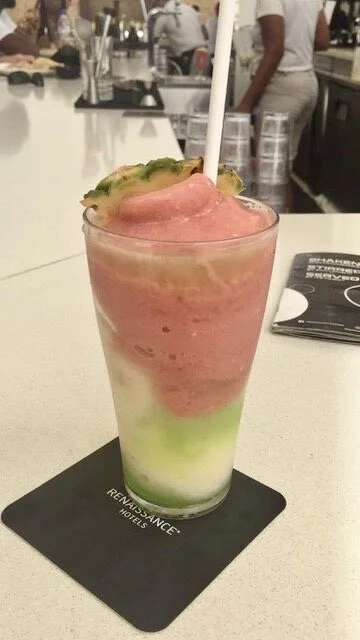 Colorful layered frozen drink in a tall glass with a straw, placed on a black coaster on the bar counter of a lively restaurant or bar.
