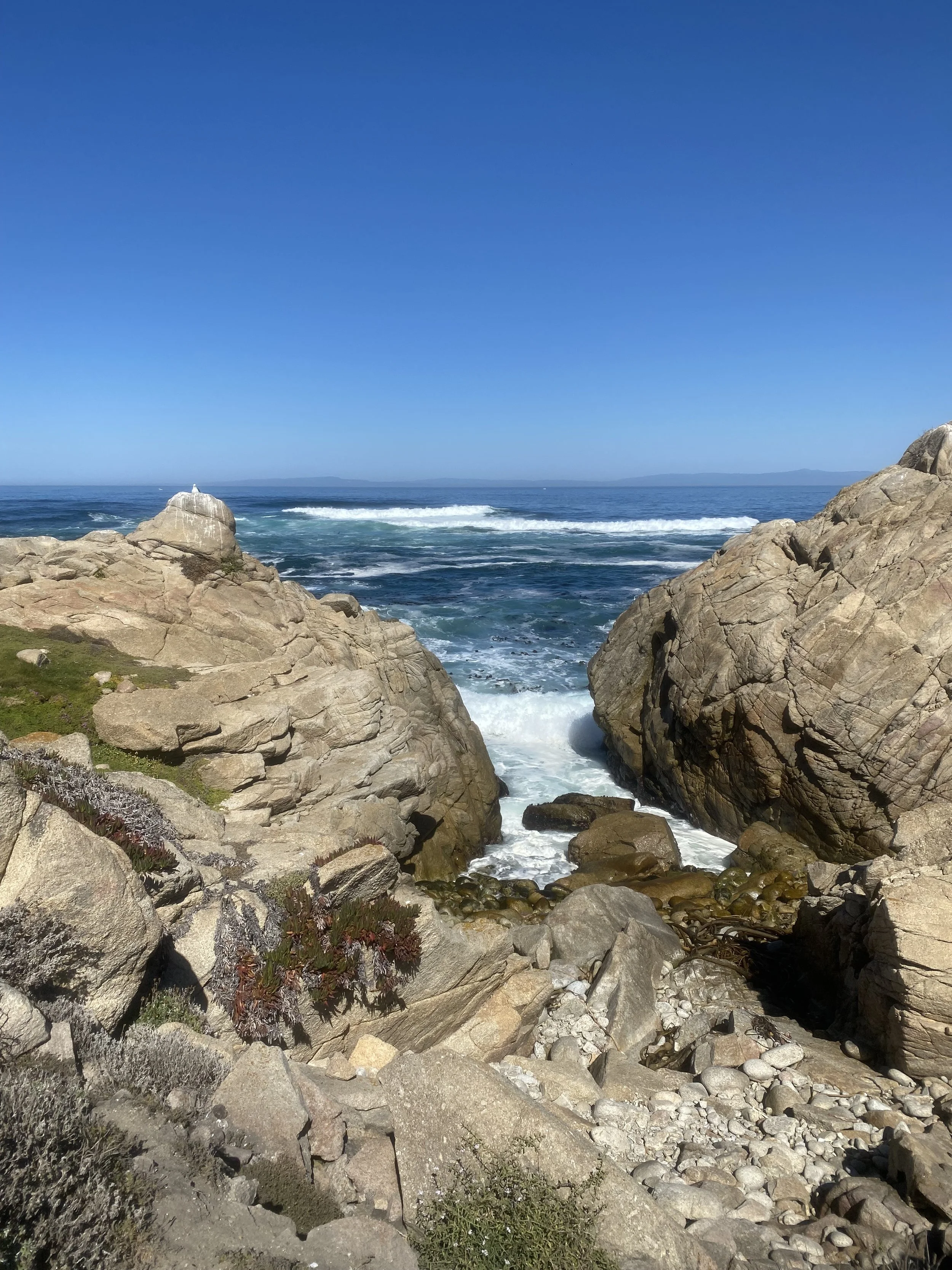 Rocky shoreline with waves crashing against large rocks, clear blue sky, distant mountains on the horizon.