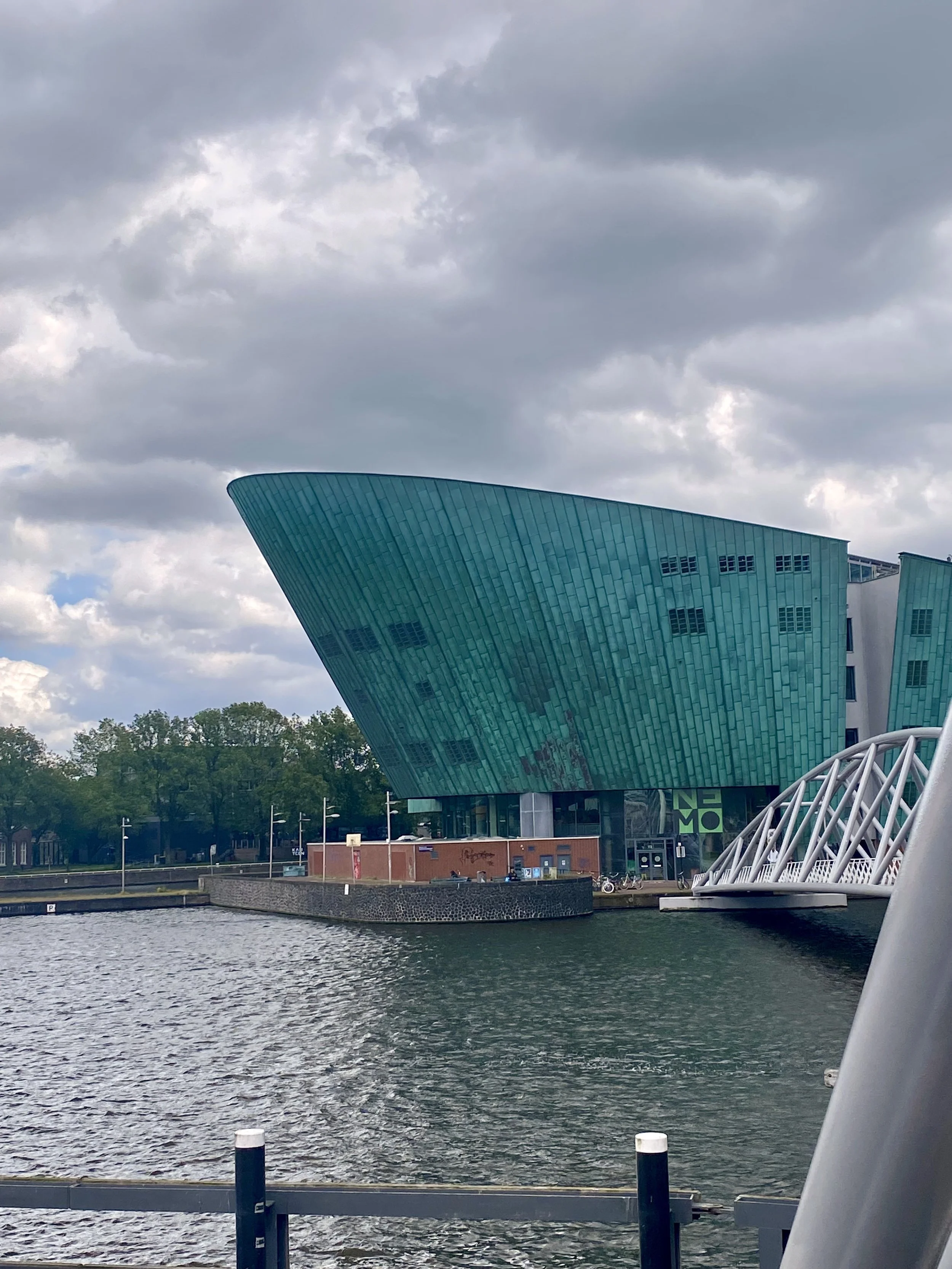 Modern building with a curved, greenish-blue facade by a body of water, overcast sky with clouds, trees and a small brick structure in the background, and a white structural element in the foreground.