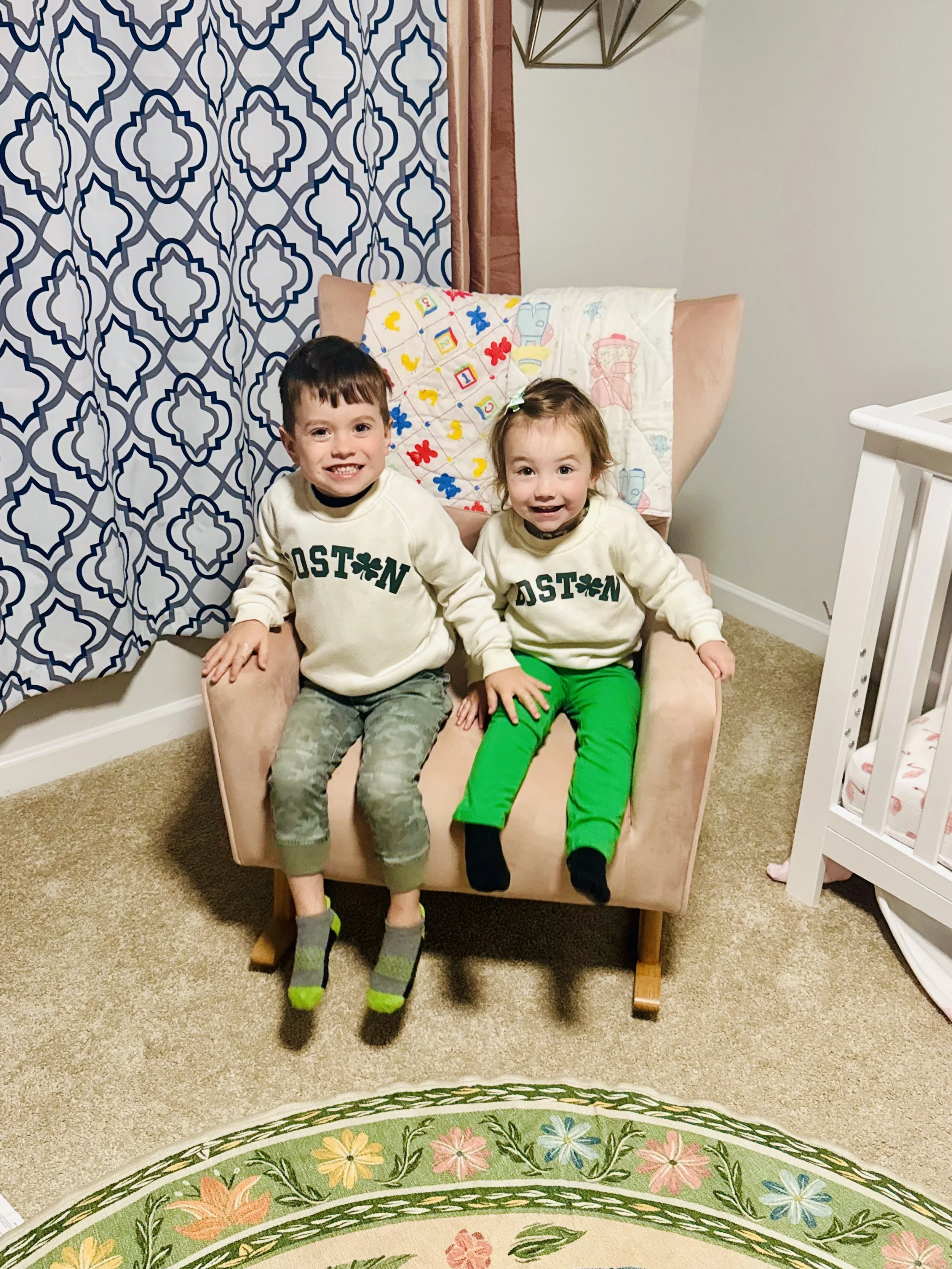 Two young children sitting on a pink armchair in a cozy room, smiling at the camera. The boy on the left wears a white sweatshirt with green lettering and camouflage pants, while the girl on the right wears a matching sweatshirt and bright green pants. The background includes patterned curtains and a quilted pillow with colorful designs, and part of a white crib is visible on the right.
