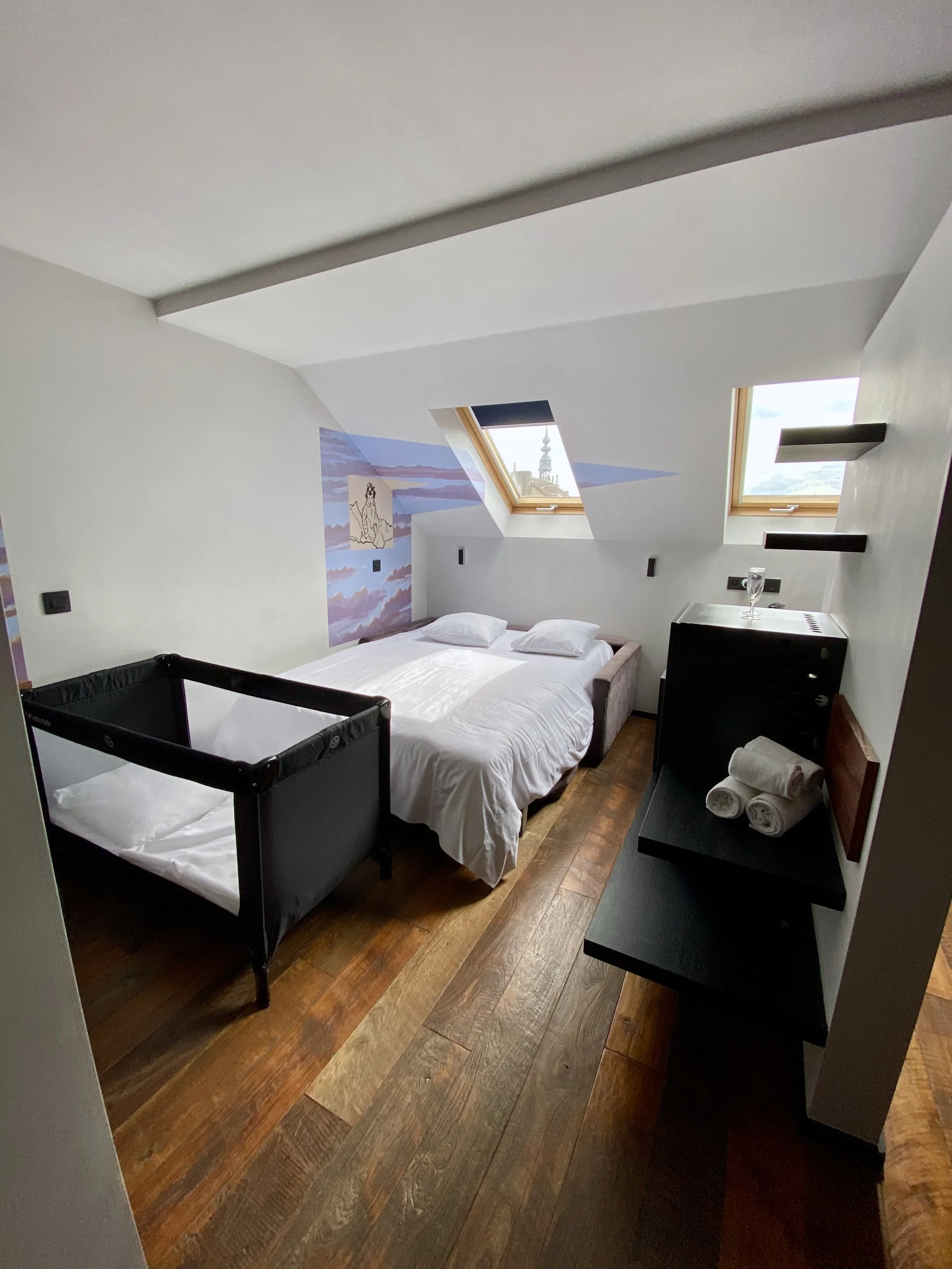 A cozy attic bedroom with a bed under skylight windows, a black side table with rolled towels, and mountain artwork on the wall.