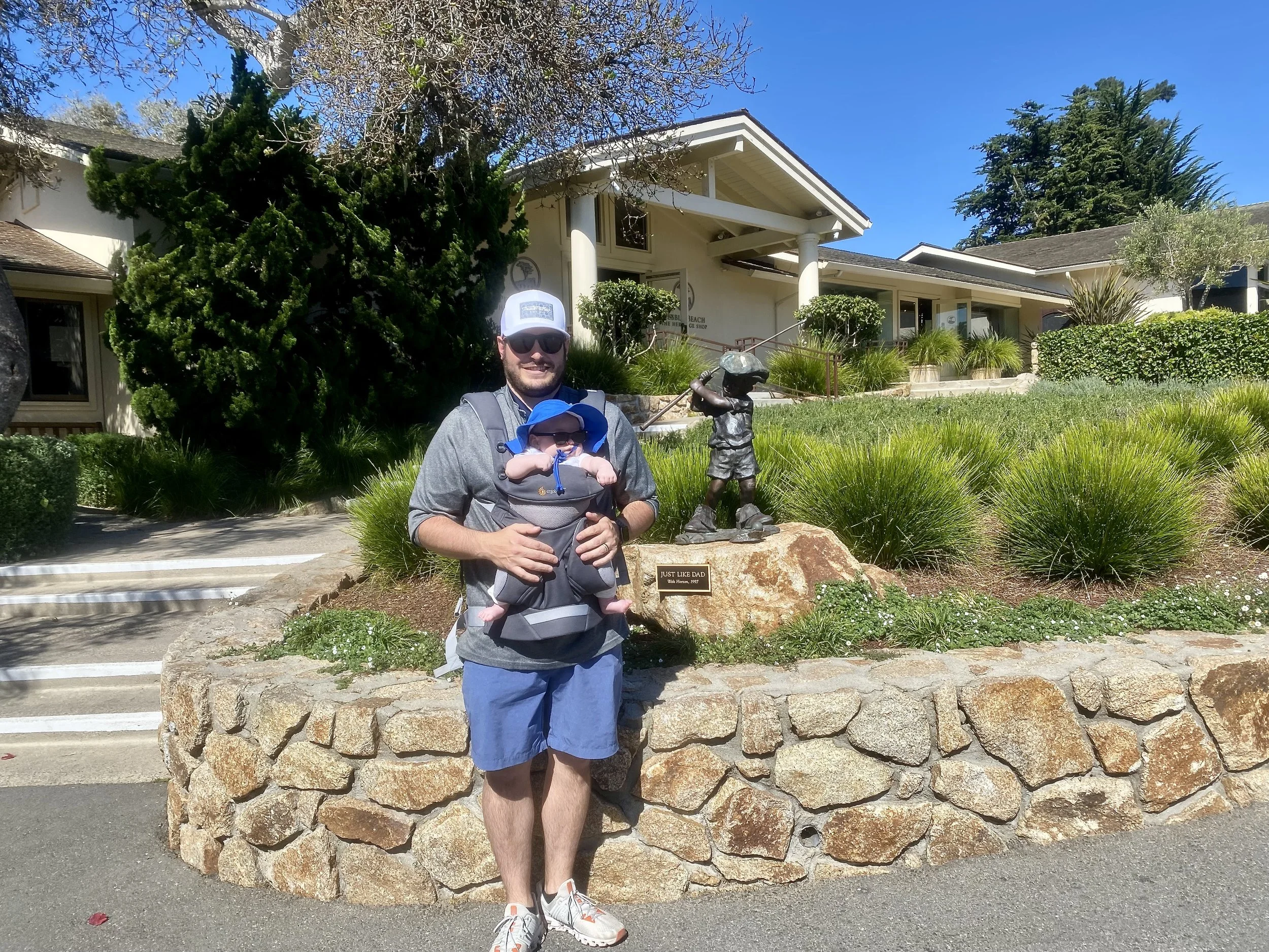 A man in sunglasses wearing a gray shirt and blue shorts is holding a baby within a front-facing carrier in front of a stone wall. They are outdoors on sunny day, with a large building and trees in the background.