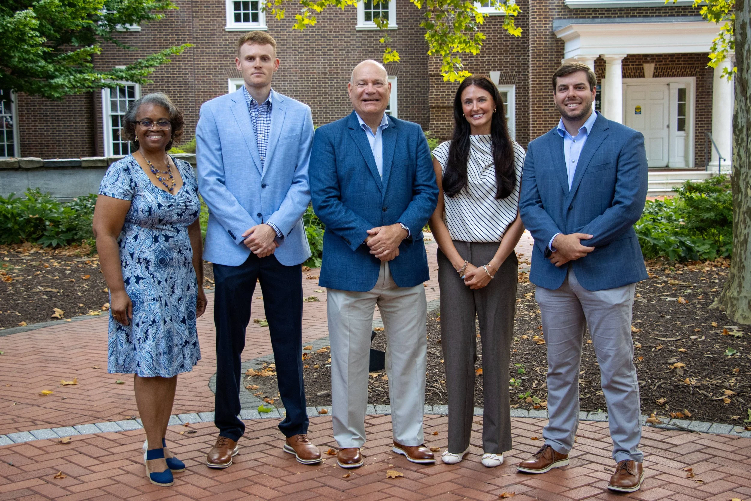 A group of five diverse professionals, three men and two women, standing outdoors in front of a brick building, smiling and dressed in business attire.