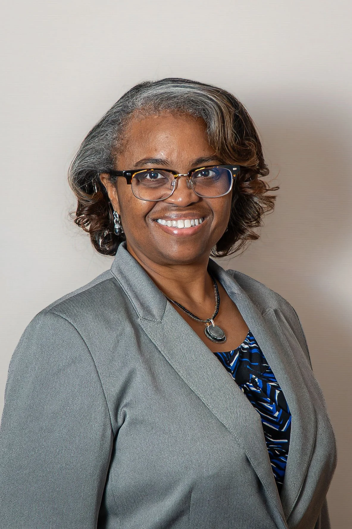 A professional Black woman with short, wavy hair, glasses, and a gray blazer, smiling at the camera against a plain light-colored background.