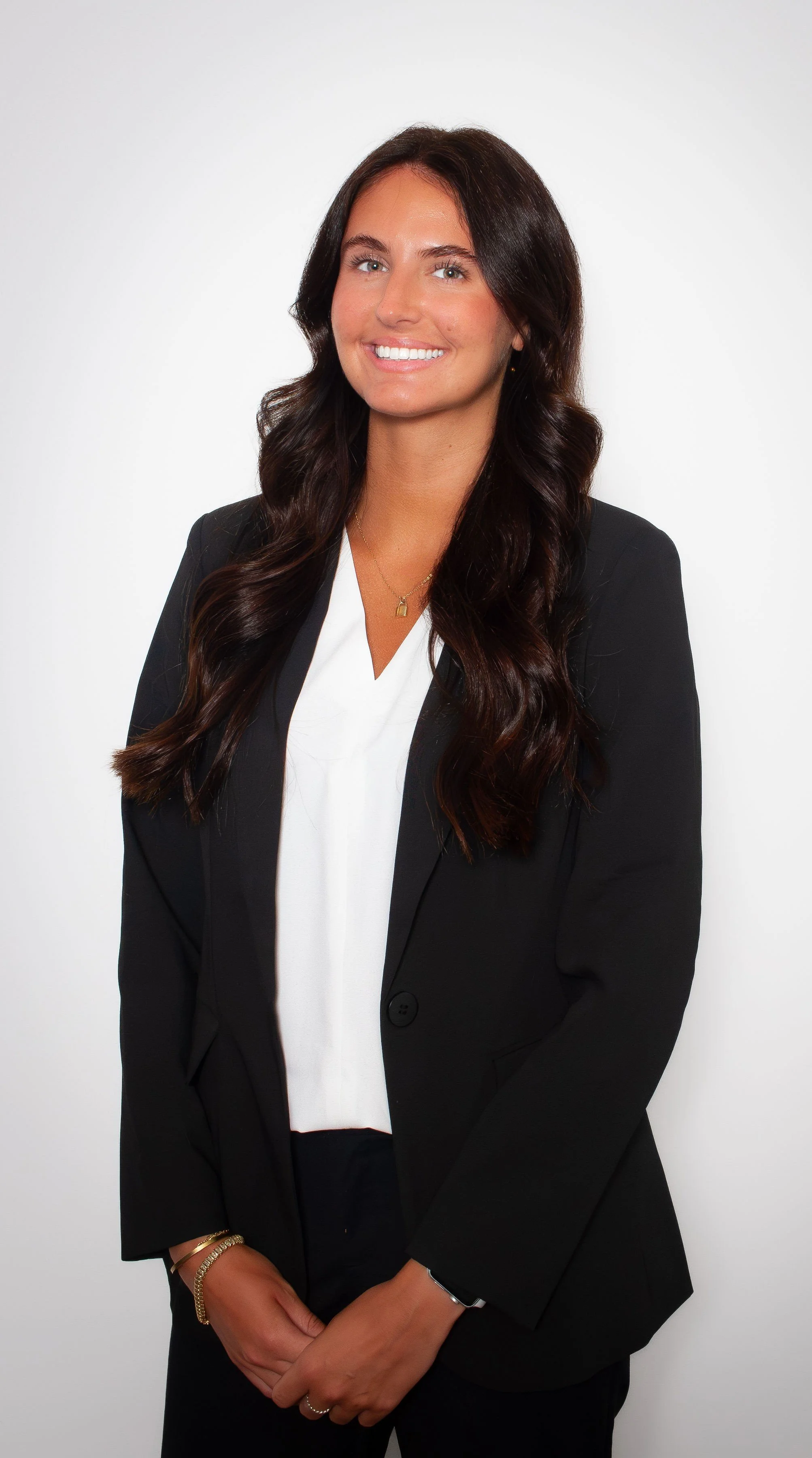 A woman with long, wavy dark brown hair, light skin, and a bright smile is dressed in a black blazer over a white blouse, accessorized with a gold necklace, bracelets, and rings, standing against a plain white background.
