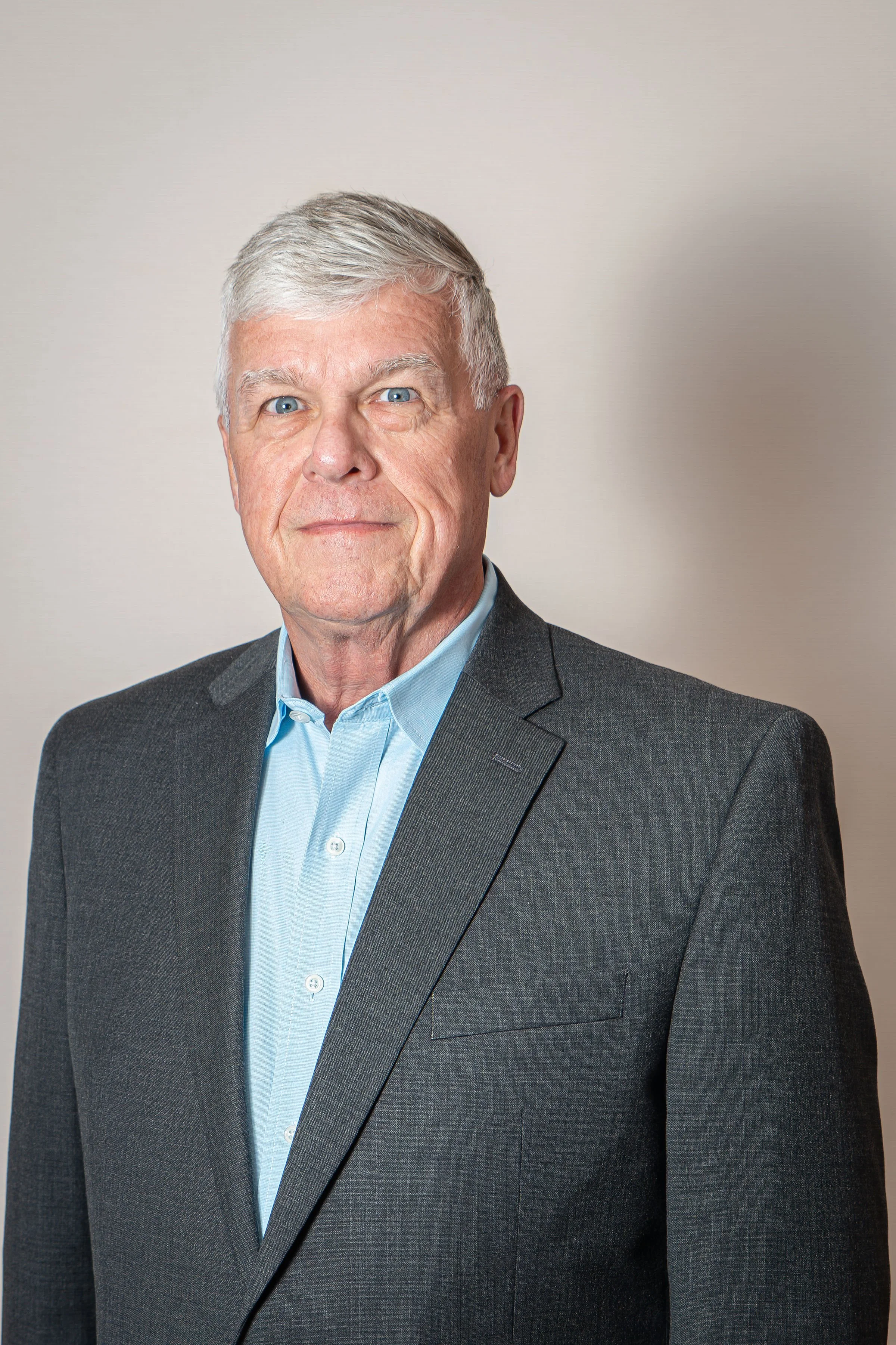 A portrait of an older man with gray hair, blue eyes, wearing a dark gray suit jacket and light blue collared shirt against a beige background.