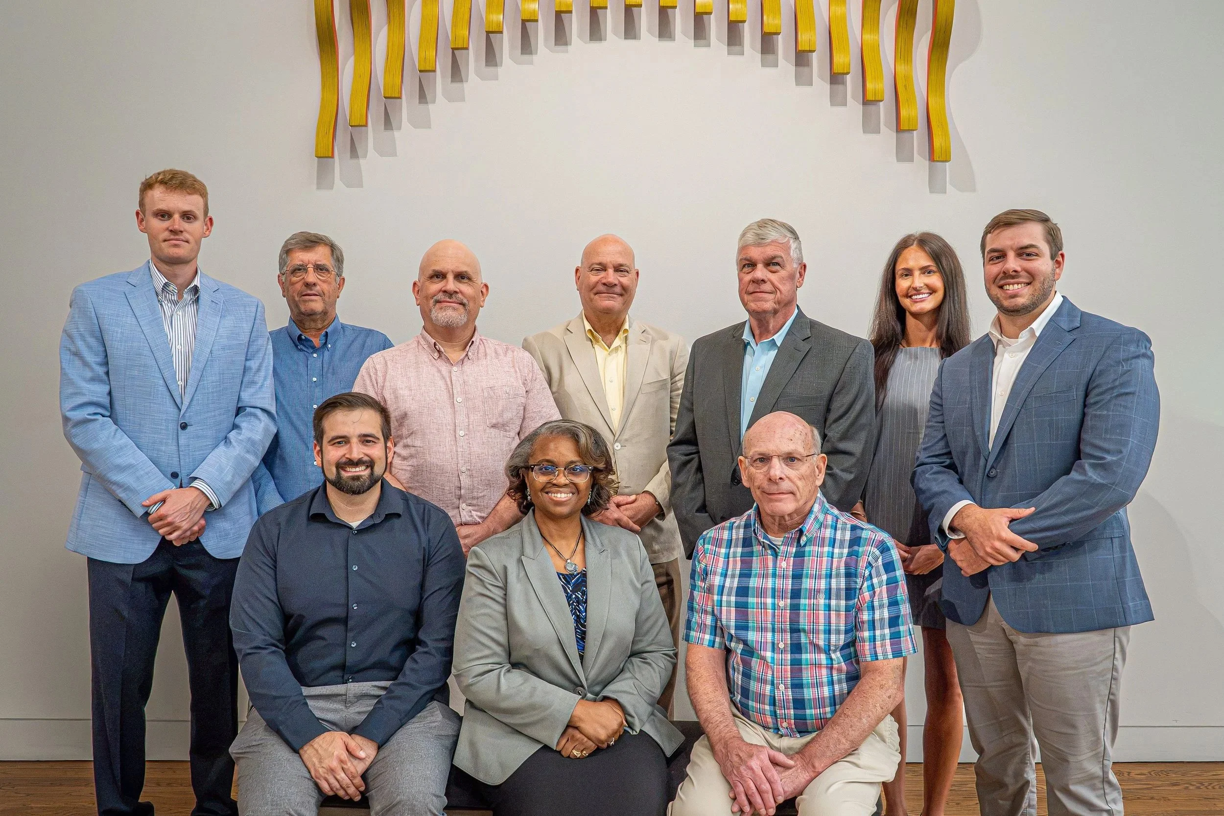 Group of men and women in business attire posing for a photo in front of a white wall with an abstract yellow wall sculpture.