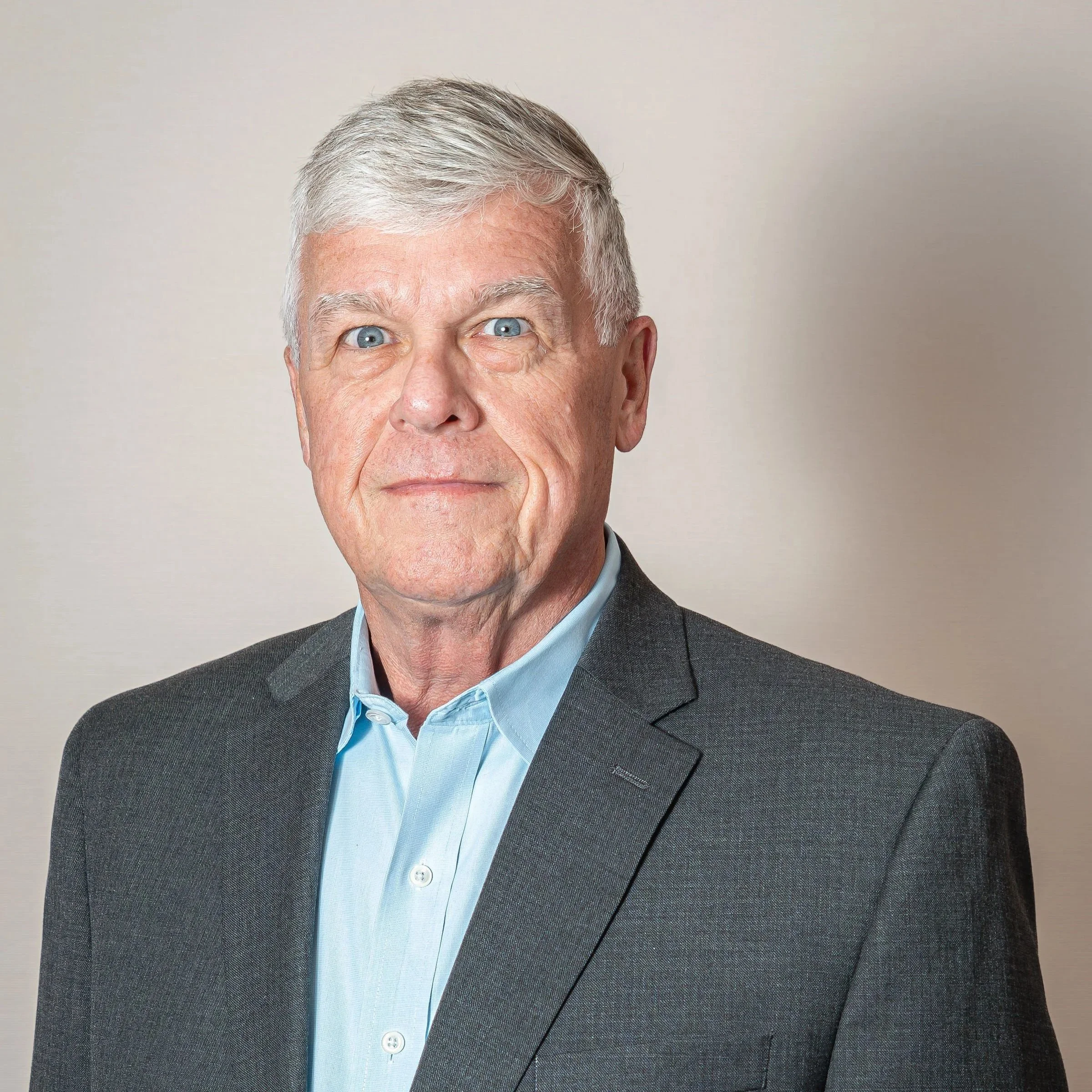 Close-up of an elderly man's face showing silver hair, blue eyes, and wearing a suit and light blue shirt, with a neutral background.