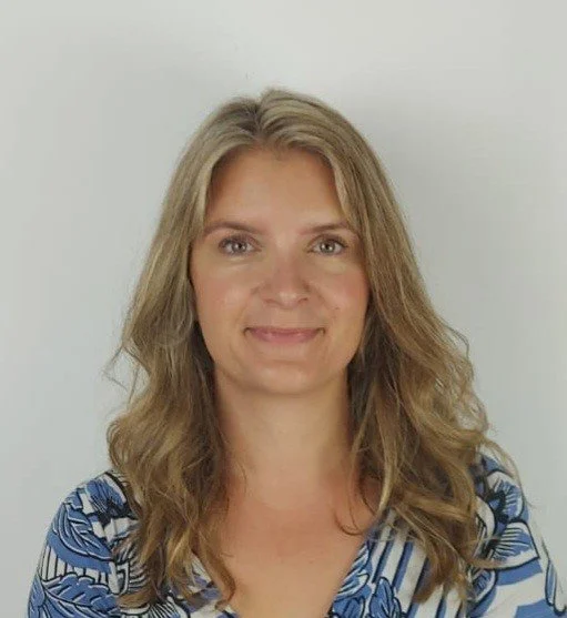Portrait of a woman with wavy blonde hair wearing a patterned blouse, smiling, against a light gray background.