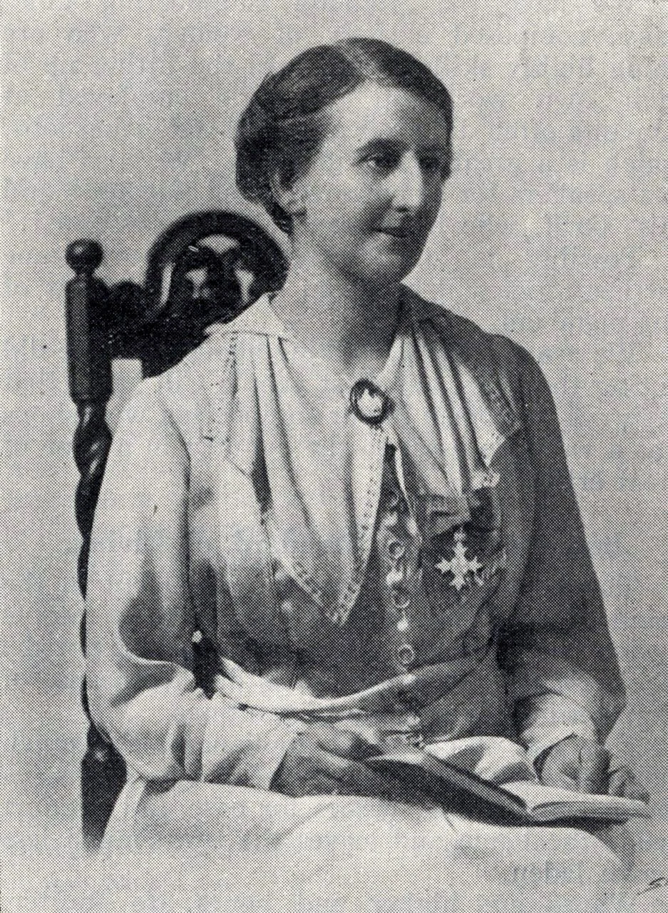 Black and white photo of a woman in a military uniform decorated with medals, sitting on a carved wooden chair, holding an open book.
