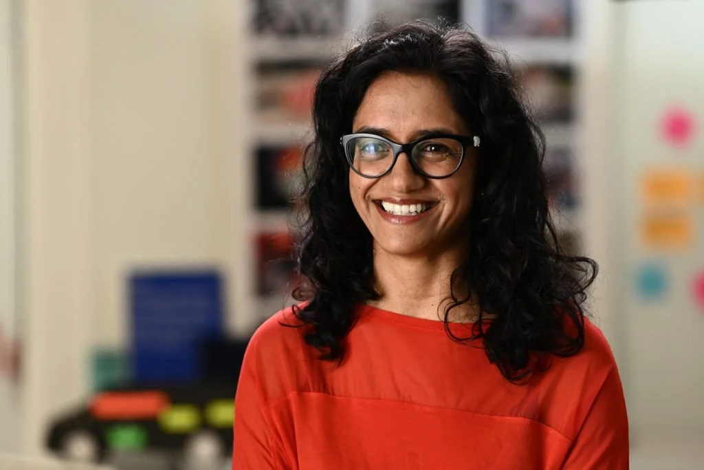 A smiling woman with curly black hair wearing glasses and a red top, standing in a room with colorful classroom posters and supplies in the background.