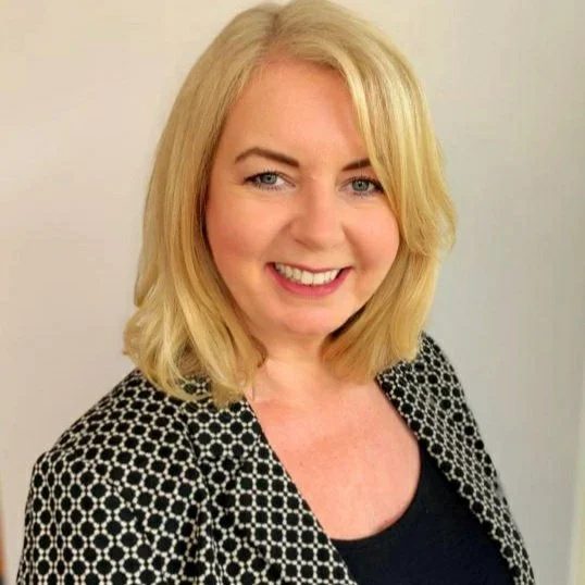 A smiling woman with shoulder-length blonde hair wearing a black top and a patterned black and white jacket.