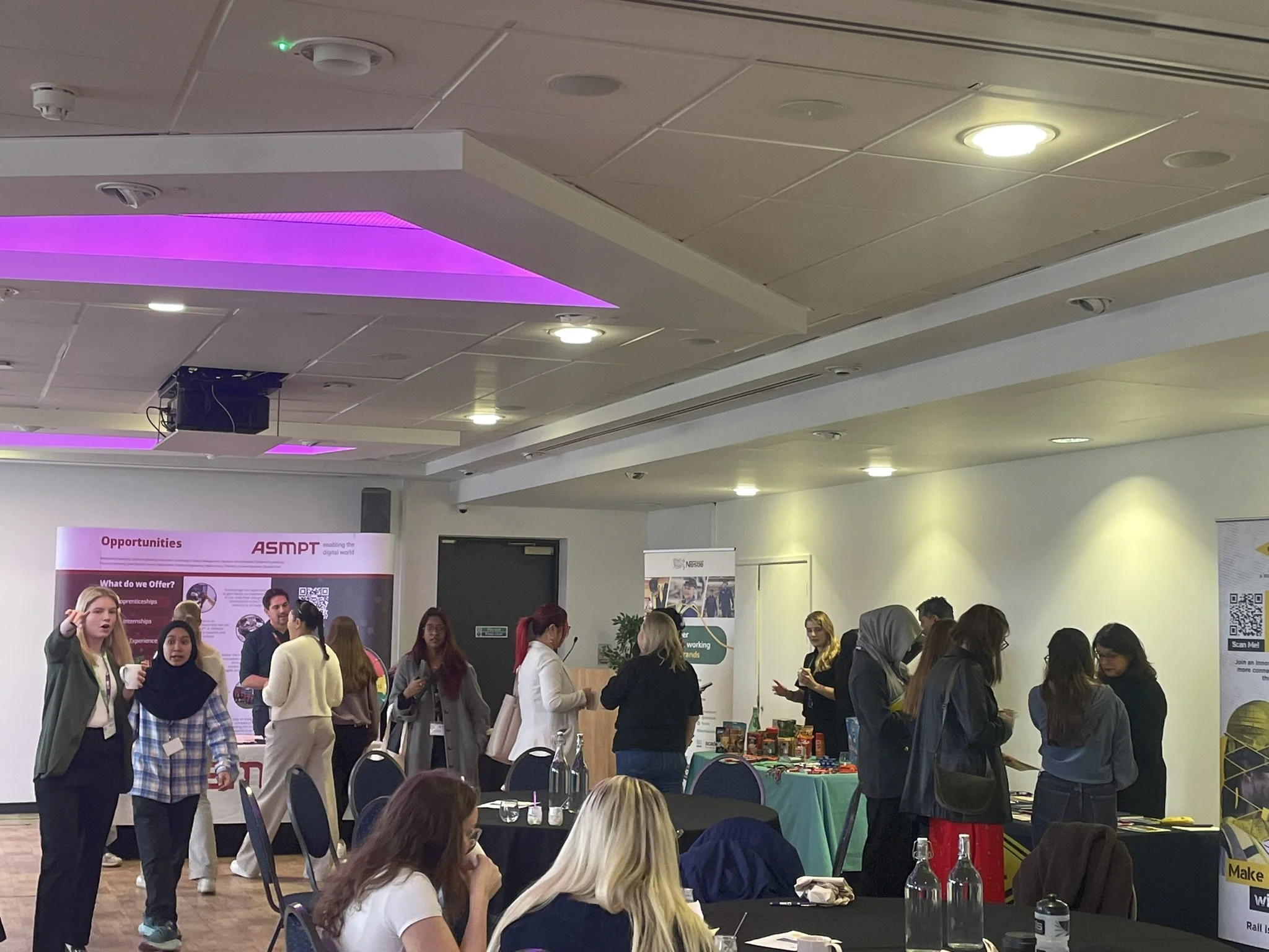 People attending a conference or exhibition in a large hall, with booths and banners, engaging in conversations and looking at displays.