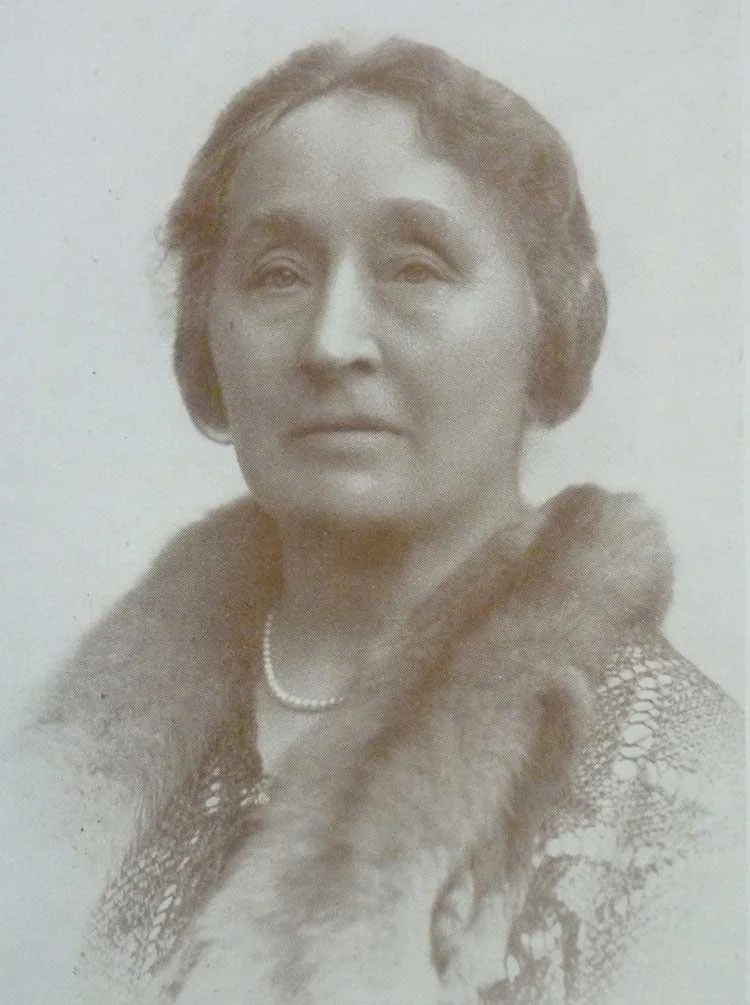 A black-and-white portrait of a woman with short hair, wearing a pearl necklace and a fur stole or collar, dressed in vintage attire.