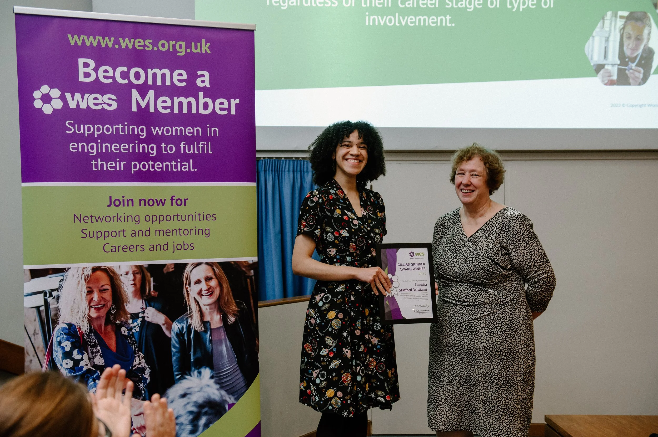 Two women standing together at an award presentation, with a large purple and green banner behind them promoting women in engineering and supporting women in the workplace.