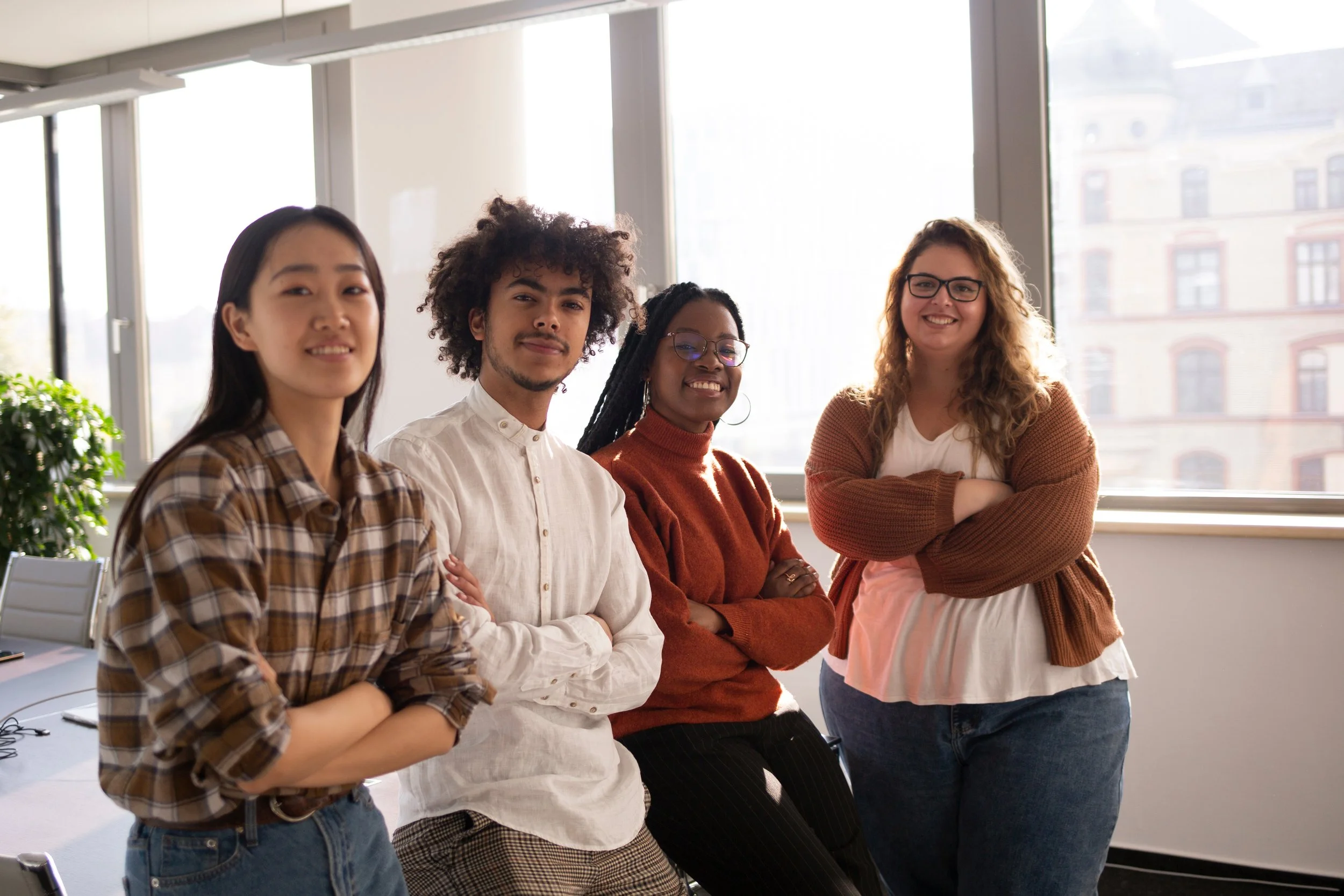 Four diverse women standing together in a bright office with large windows, smiling and looking at the camera.