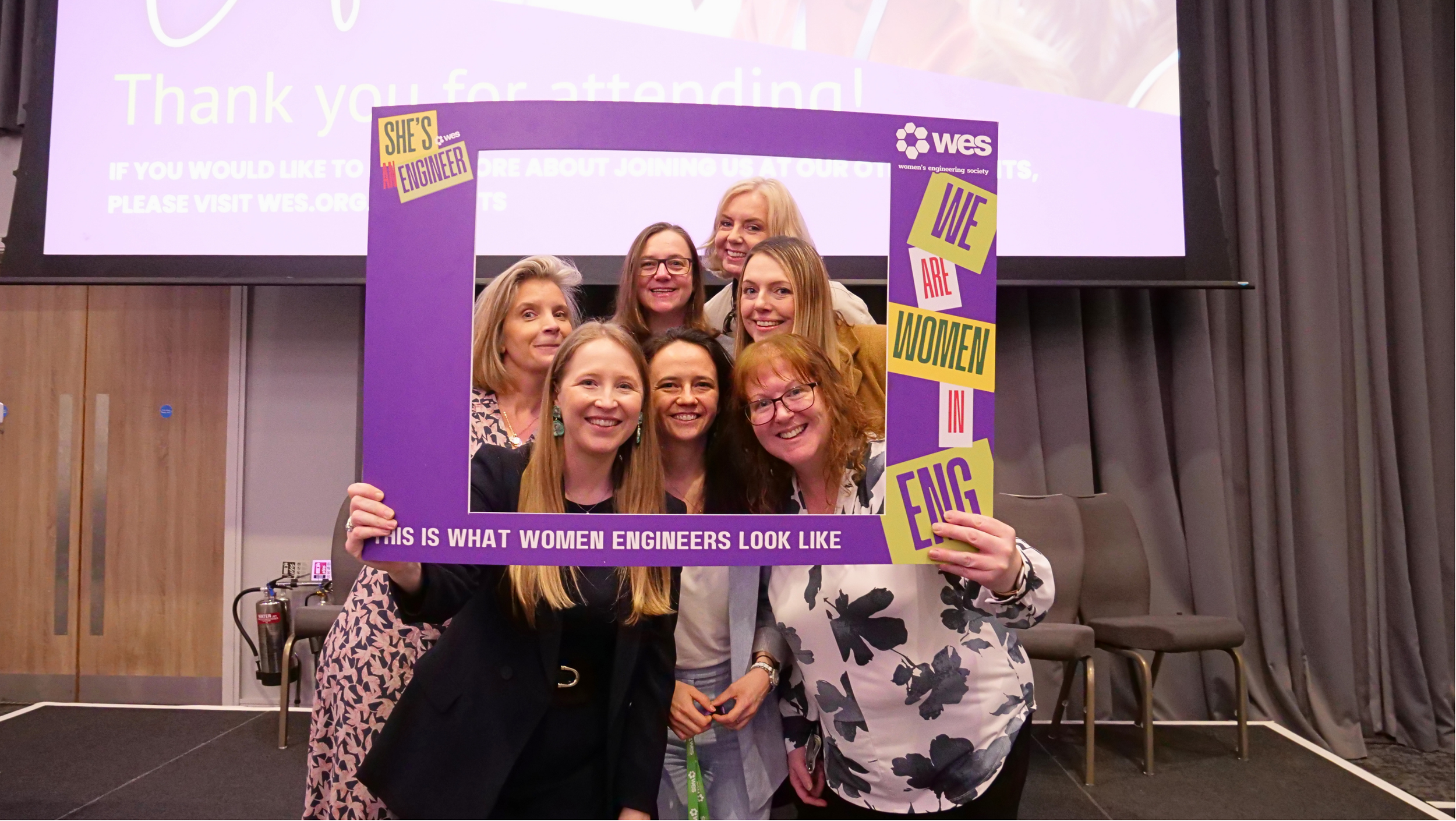 A group of women holding a large purple frame with the word 'WES' and its logo, stating 'Women’s Engineering Society,' with the phrase 'This is what women engineers look like' at the bottom. The women are smiling and standing together at an event.