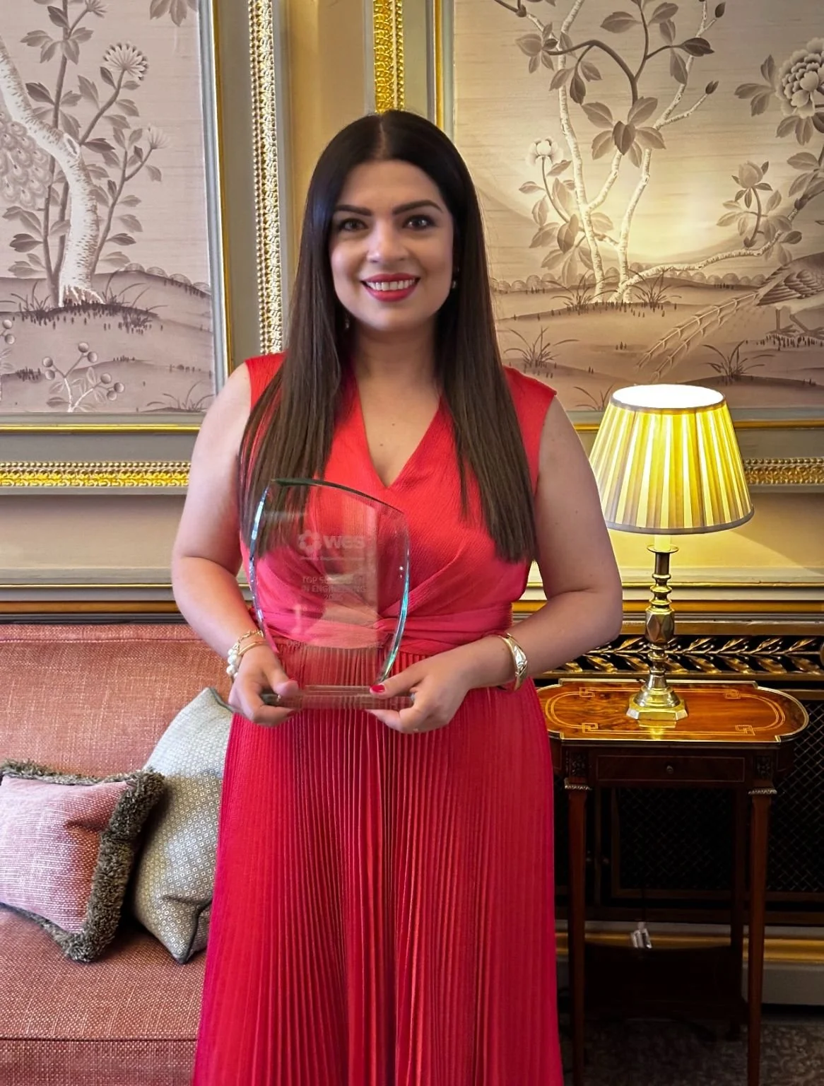 A woman with long dark hair wearing a red dress holding an award trophy in a luxurious room with gold framed artwork and a yellow striped lampshade.