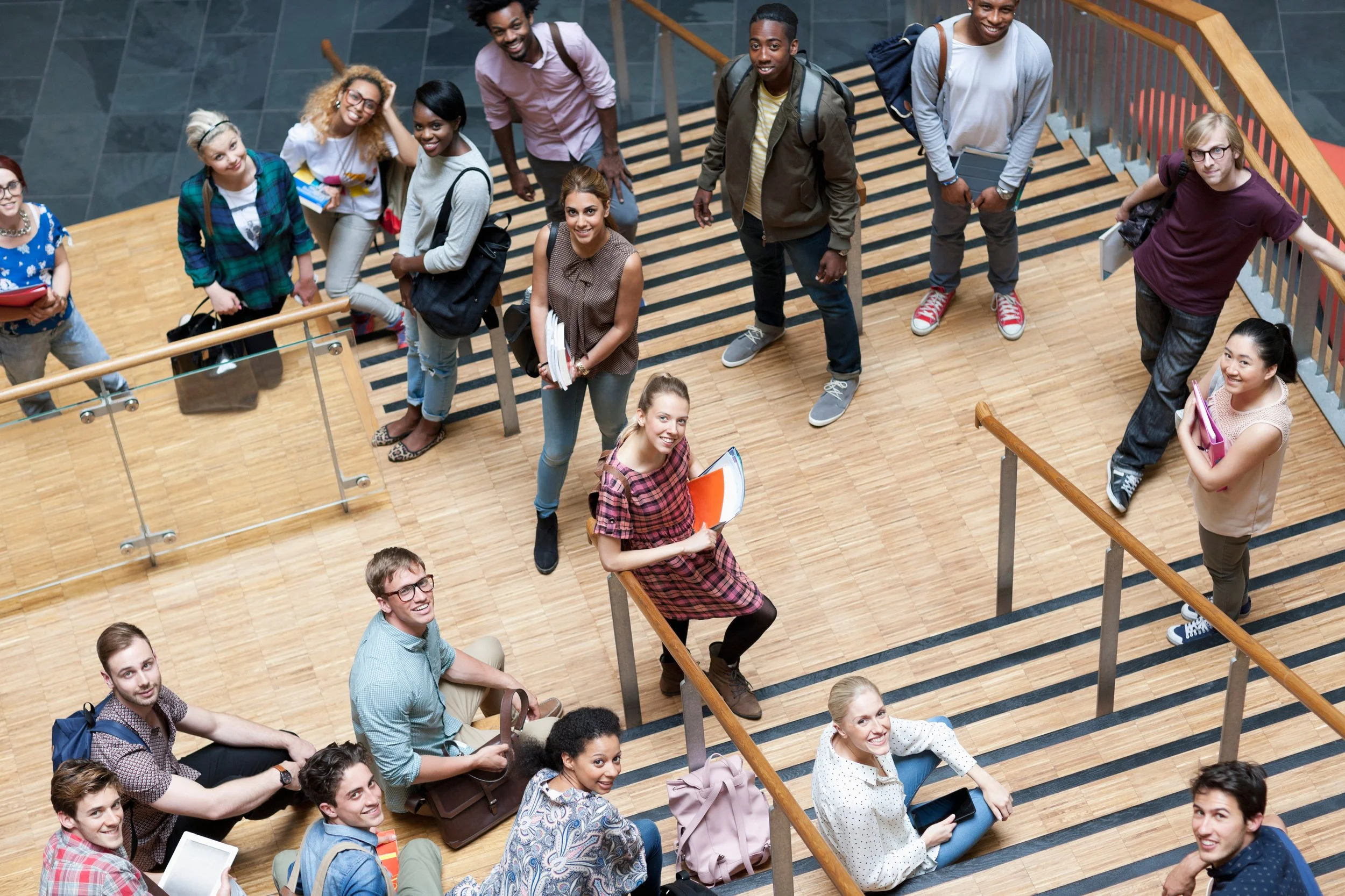 Group of diverse college students standing on a wooden staircase in a modern building, smiling and looking up at the camera, carrying backpacks and books.