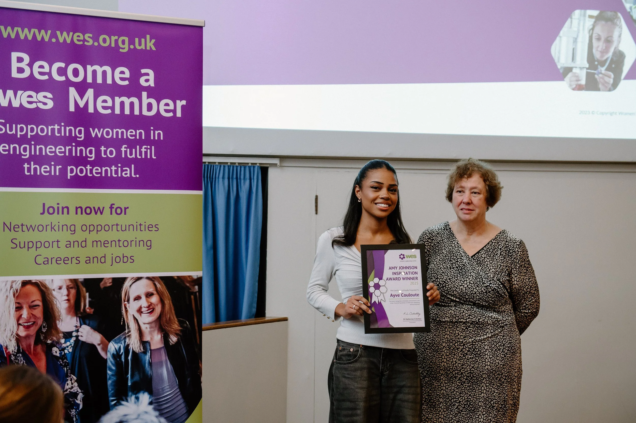 A woman receiving an award from another woman at an event. The woman on the left is holding a framed certificate, and the woman on the right is wearing a patterned dress. There is a purple and green banner to the left with text about supporting women in engineering.