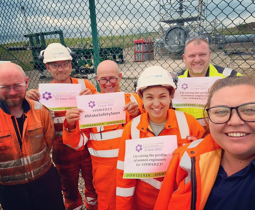 Group of six women and men in safety gear holding signs at an engineering day event, with a fenced industrial site and large industrial equipment in the background.