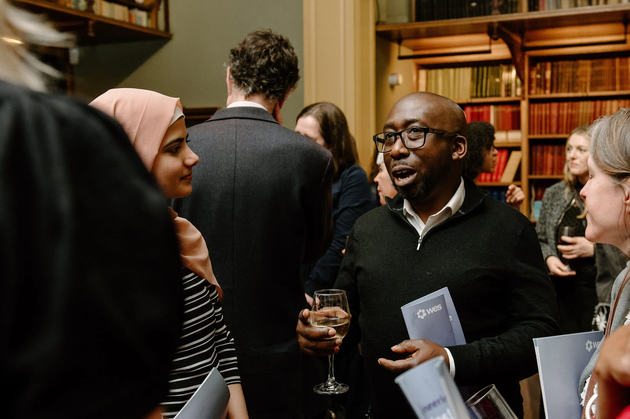 People engaged in conversation at a social event in a room with bookshelves.