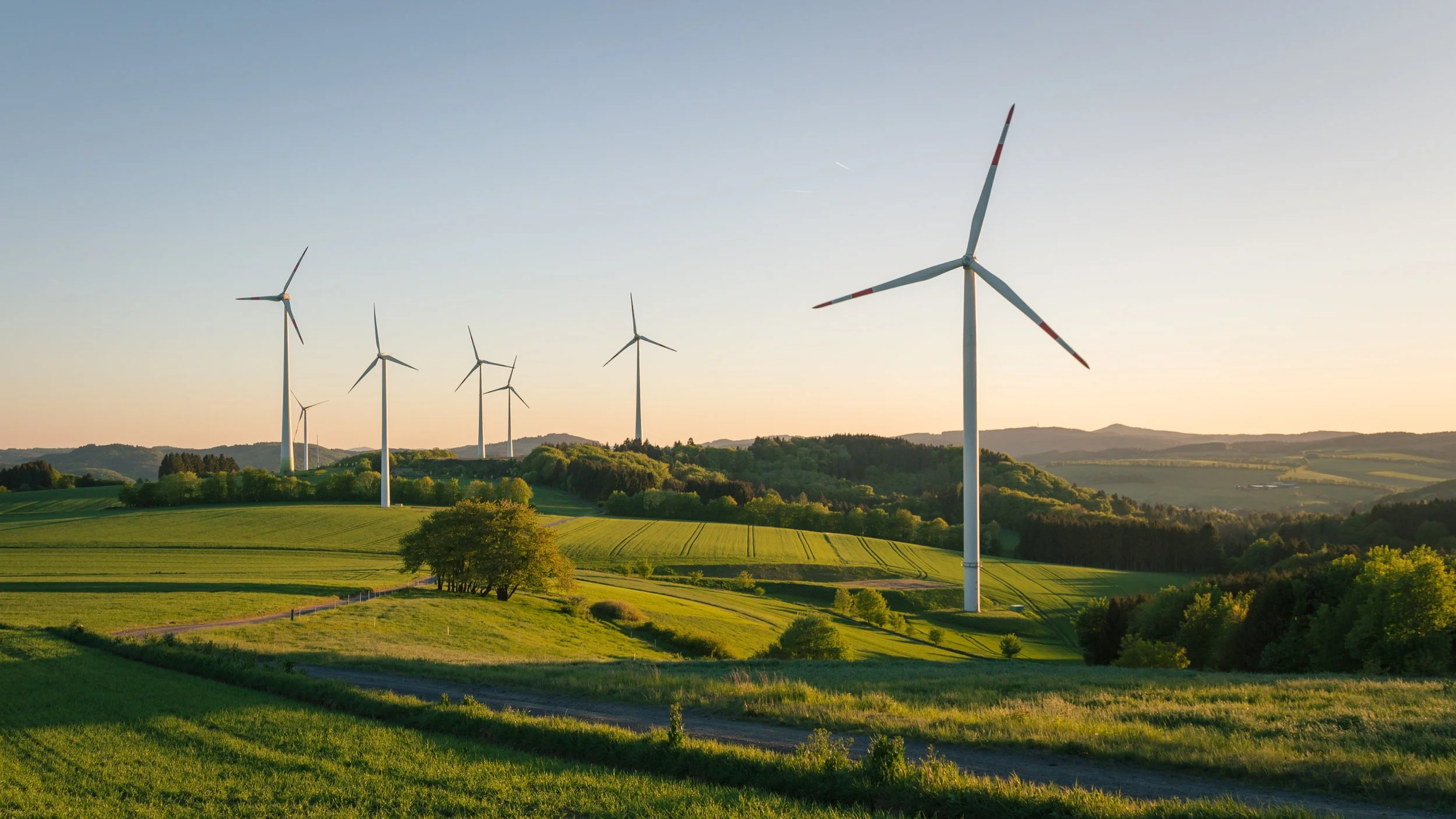Rolling green fields with multiple wind turbines on a hill, under a clear blue sky with the sun setting in the distance.