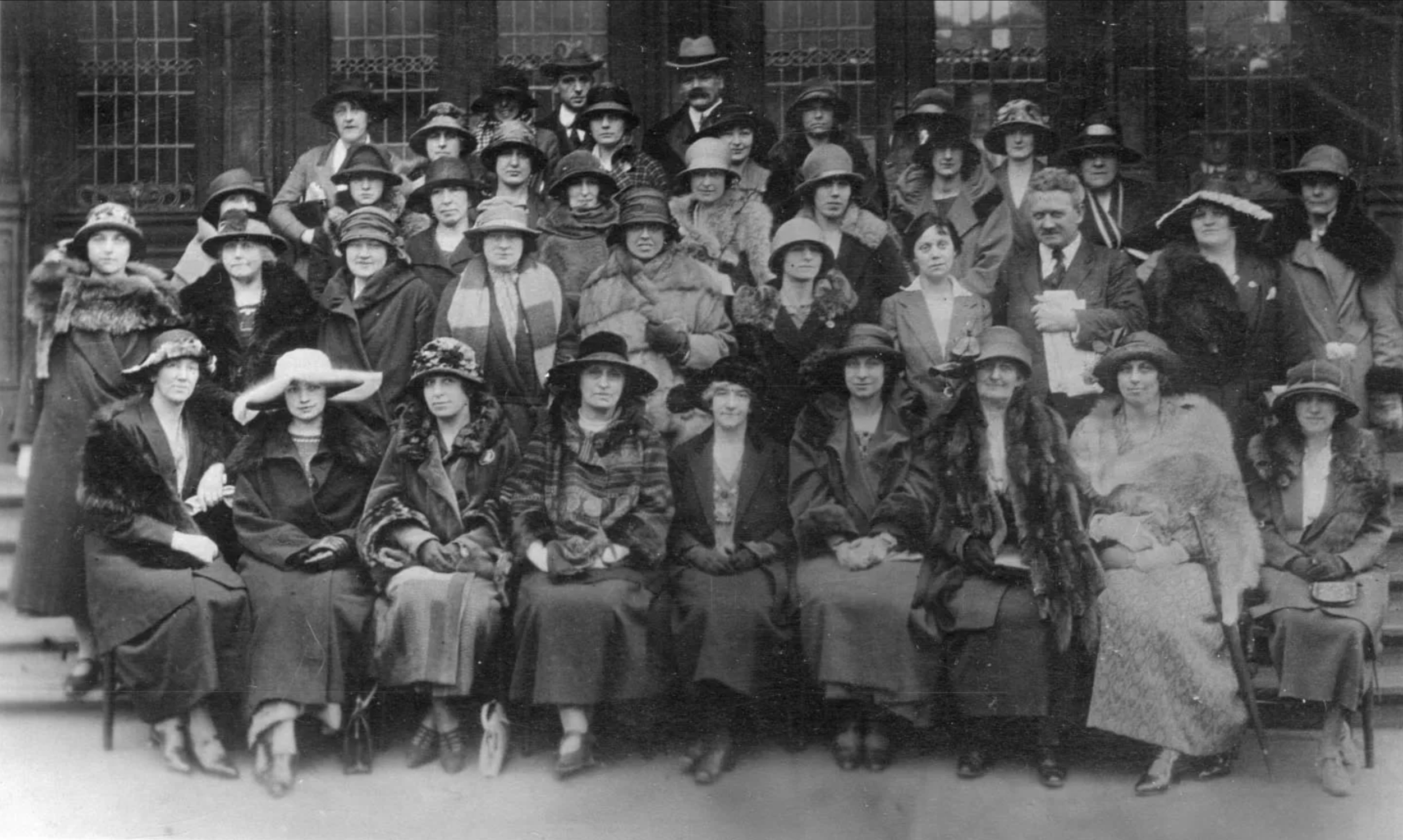 Group portrait of women and men dressed in early 20th-century clothing, including hats and coats, standing and sitting in front of a building with decorative ironwork.