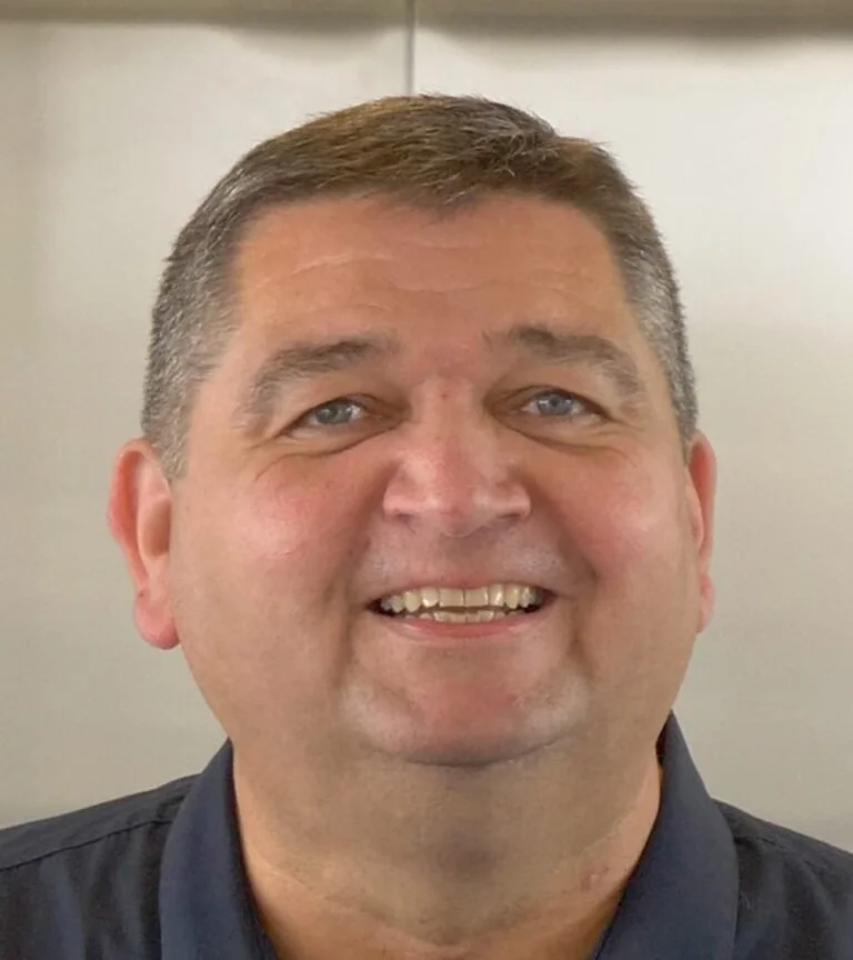 A smiling middle-aged man with short brown hair, blue eyes, and a fair complexion, wearing a dark collared shirt.