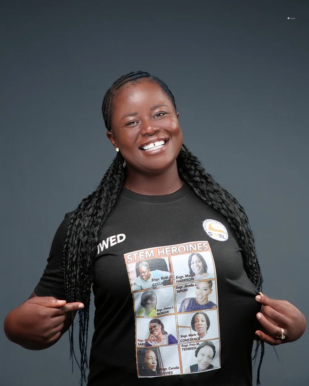 A smiling woman with braided hair pointing to her chest, wearing a black T-shirt with a collage of women labeled 'STEM HEROINES'.