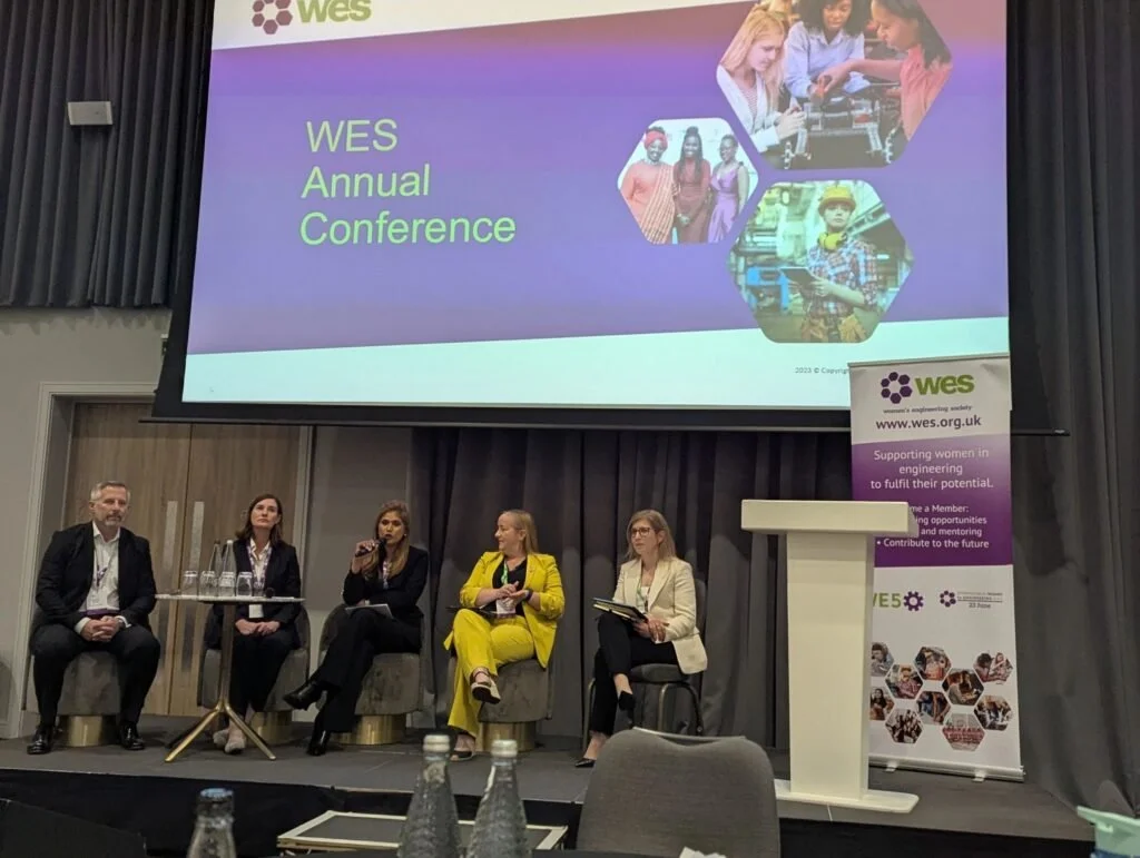 A panel of five people seated on stage at the WES Annual Conference, with a large screen above displaying the conference's name and logo, and a standing banner to the right supporting women in engineering.