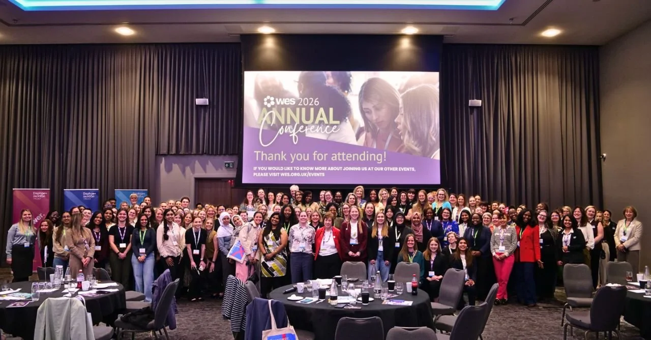 Group photo of diverse women at a conference, with a large screen behind them displaying conference details and a thank you message.