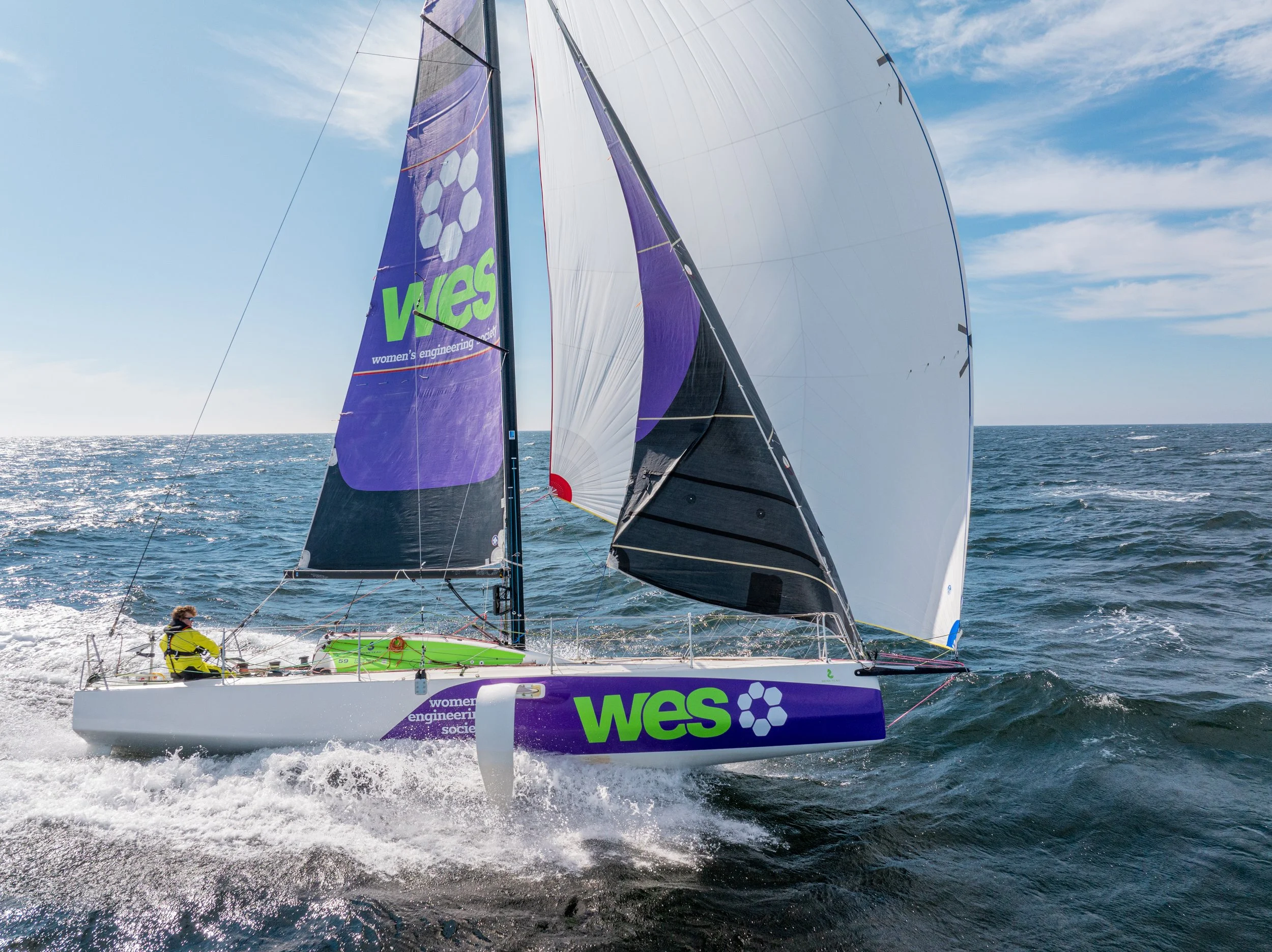 A sailboat with large purple and white sails on the open ocean, carrying a crew member wearing a yellow life jacket. The sailboat features logos for WES and women’s engineering society, and the ocean is glistening under a partly cloudy sky.