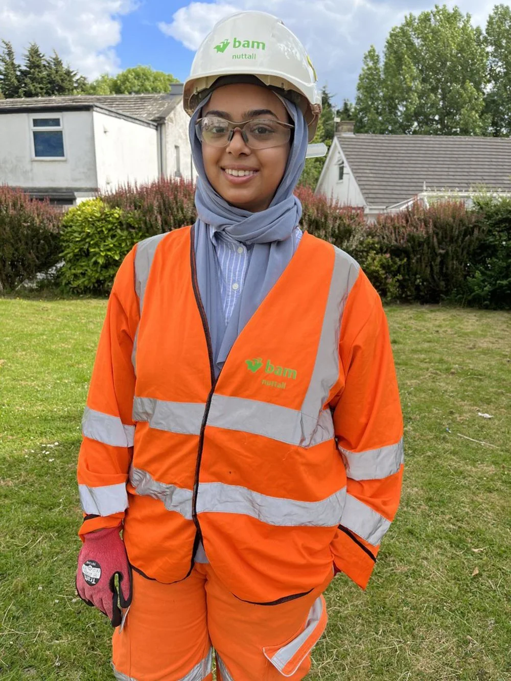 A woman wearing safety gear, including an orange high-visibility jacket, a helmet with a logo, safety glasses, a headscarf, and work gloves, standing outdoors with houses and greenery in the background.