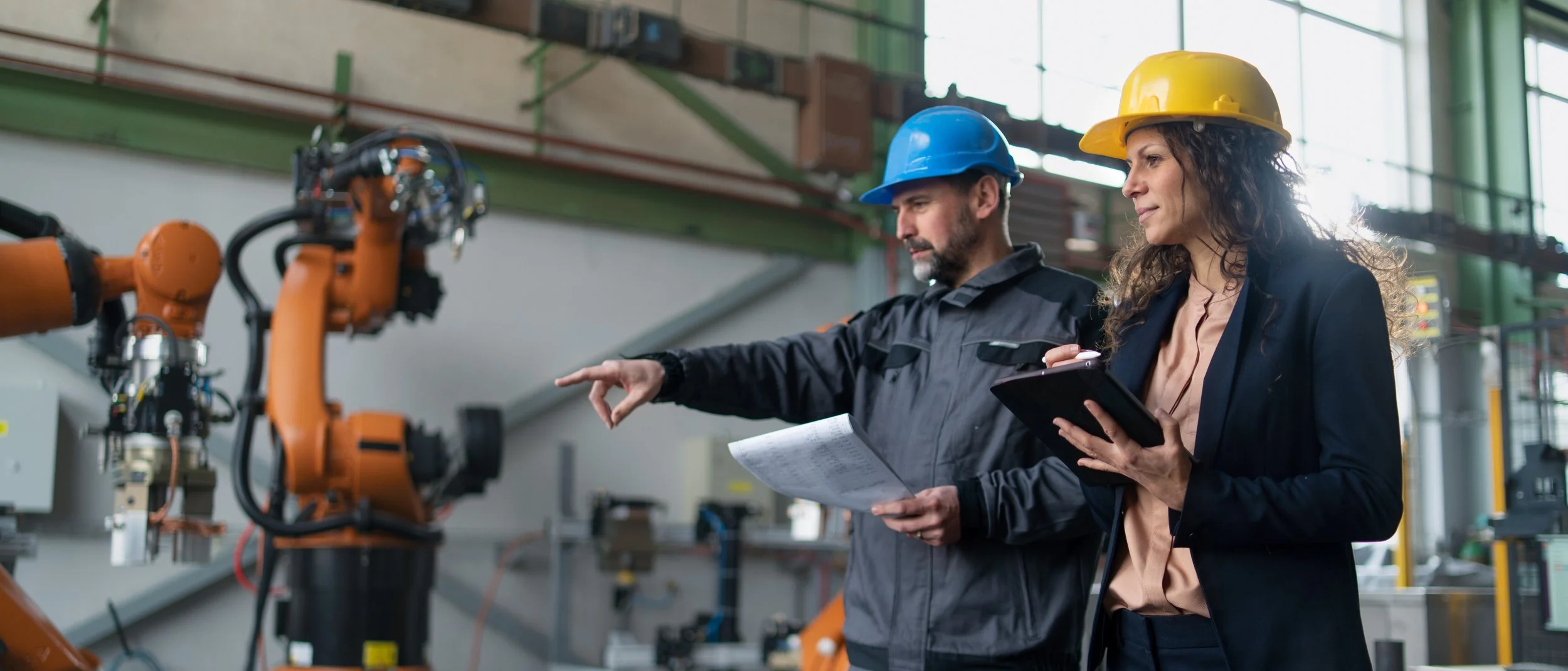 Two factory workers, a man and a woman, are inspecting a large robotic arm in an industrial setting. The man is pointing at the robot and holding a notepad, while the woman is holding a tablet and wearing a yellow safety helmet. Both are dressed in safety gear and work uniforms.