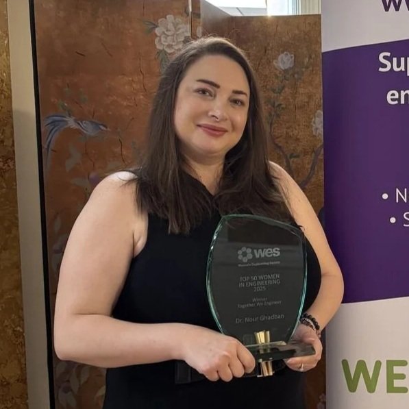 A woman with long dark hair, wearing a black sleeveless top, holding an award plaque with both hands, standing next to a purple and white banner.