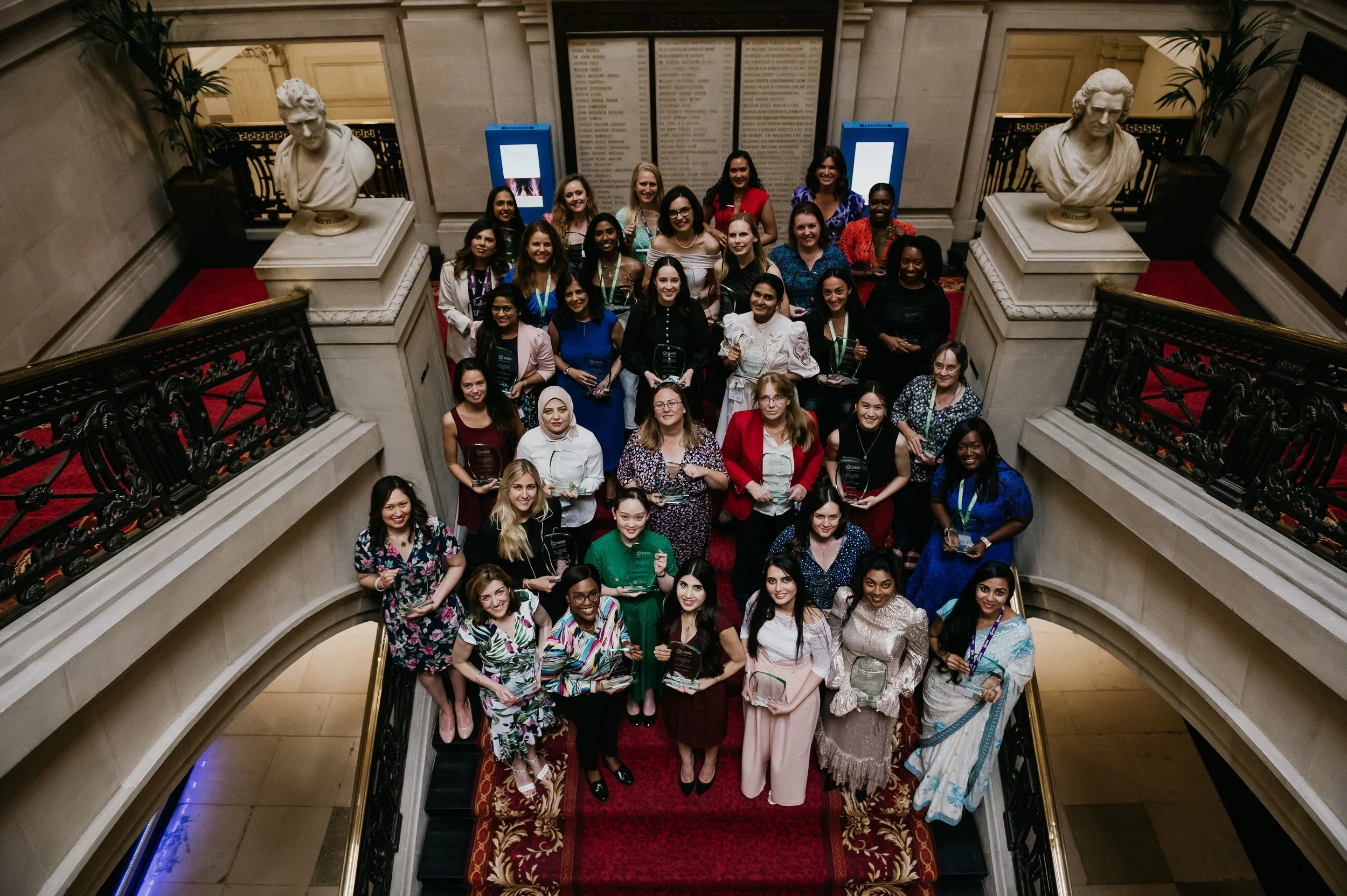 Group of women posing on a red-carpeted staircase inside a grand building, with marble busts and informational signs in the background.