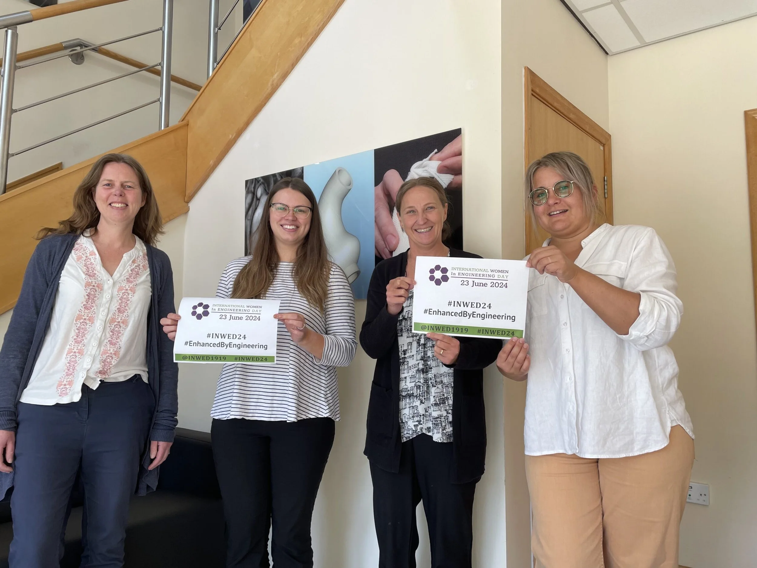 Four women standing indoors posing for a photo, holding signs celebrating International Women in Engineering Day on June 23, 2024, with a staircase and wall posters in the background.